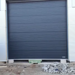 Zamknięta, segmentowa, antracytowa brama garażowa w nowym budynku przemysłowym z widocznymi fragmentami betonu i gruzu na ziemi przed nią.