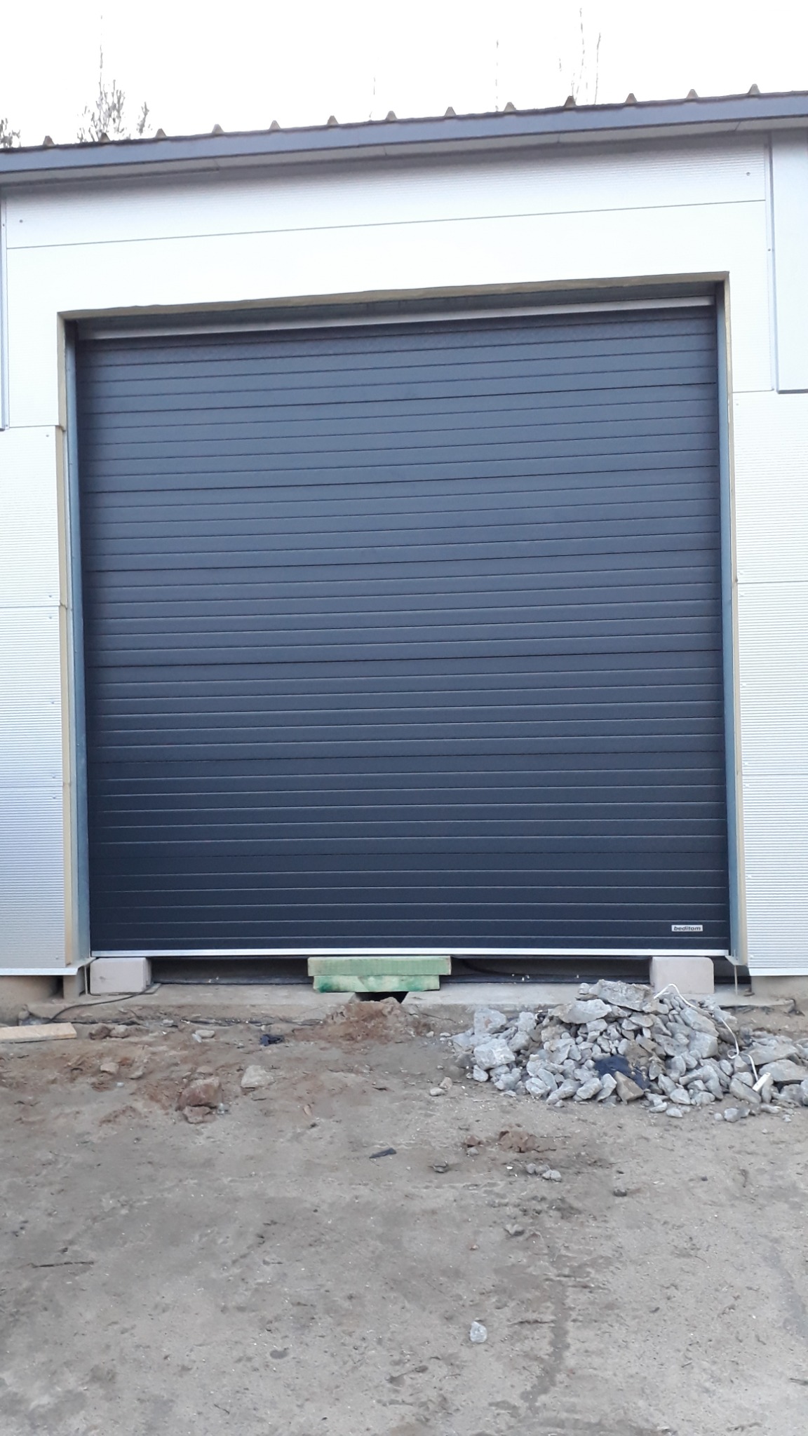 Zamknięta, segmentowa, antracytowa brama garażowa w nowym budynku przemysłowym z widocznymi fragmentami betonu i gruzu na ziemi przed nią.
