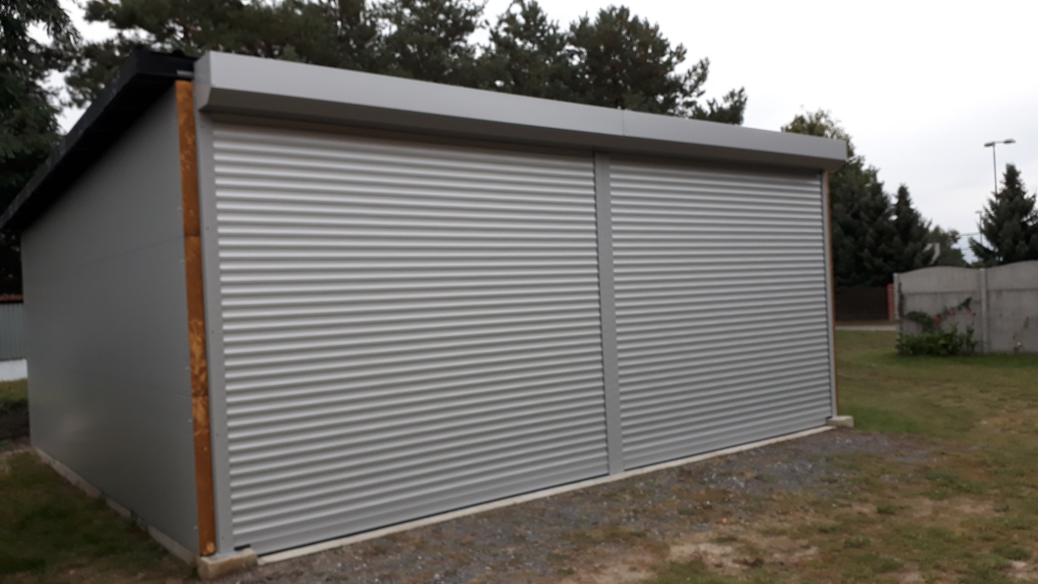 Szary garaż blaszany z dwoma segmentami rolowanej osłony, widok z boku na tle trawy i drzew.