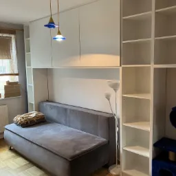 Szara sofa z tapicerowanymi bokami i kocem, wkomponowana w białą zabudowę meblową z szafkami górnymi i regałami po bokach, oświetlona wiszącą lampą z niebieskimi kloszami.