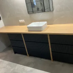 Nowoczesna szafka łazienkowa z blatem drewnianym i czarnymi frontami z ryflowaniem, umywalka nablatowa, lustro w czarnej ramie. Elementy wykończenia w odcieniach beżu i szarości.