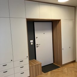 MNW PW FL STUDIO - Białe szafy wnękowe z drewnianymi akcentami otaczają wejście z drzwiami. Widoczna drewniana podłoga w jodełkę i siedzisko. Minimalistyczny design, ciepłe oświetlenie.