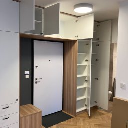 MNW PW FL STUDIO - Biała szafa wnękowa z drewnianymi akcentami, wkomponowana we wnękę przy drzwiach wejściowych. Otwarta szafka z półkami i szuflady z czarnymi uchwytami.