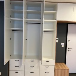 MNW PW FL STUDIO - Biała szafa wnękowa z otwartymi drzwiami, widoczne półki i drążki na ubrania, dolne szuflady z czarnymi uchwytami, drewnopodobna podstawa i siedzisko obok.