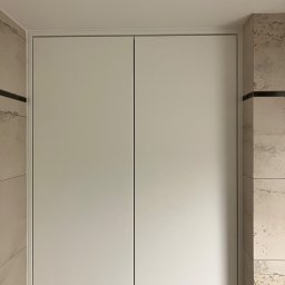 MNW PW FL STUDIO - Biała szafka wnękowa z dwoma drzwiami, zamontowana w łazience z dekoracyjnymi płytkami. Widoczne przewody elektryczne u dołu szafki.