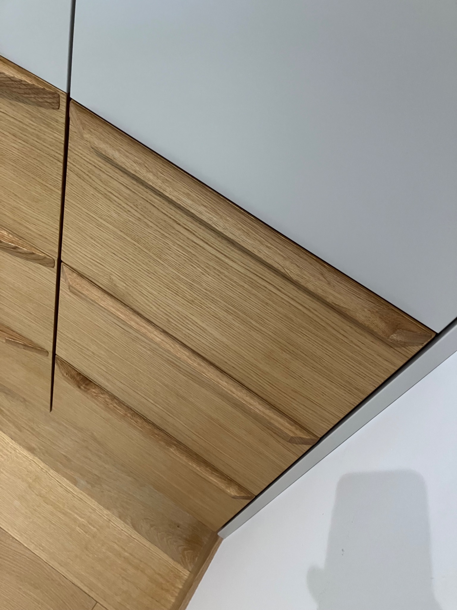 Drewniane fronty szafy z subtelnymi uchwytami frezowanymi, połączone z gładkimi, szarymi powierzchniami. Ujęcie z bliska, ukazujące detale wykończenia i strukturę drewna.