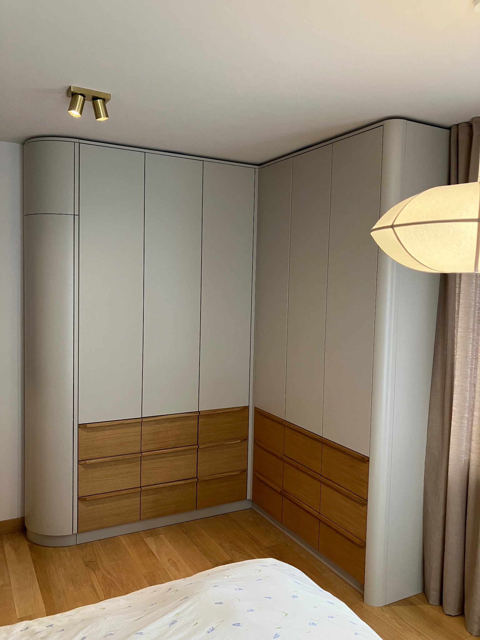 Jasnoszara szafa narożna z drewnianymi szufladami, zaokrąglone krawędzie, oświetlenie punktowe i lampa wisząca. Parkiet na podłodze, widok z dołu.