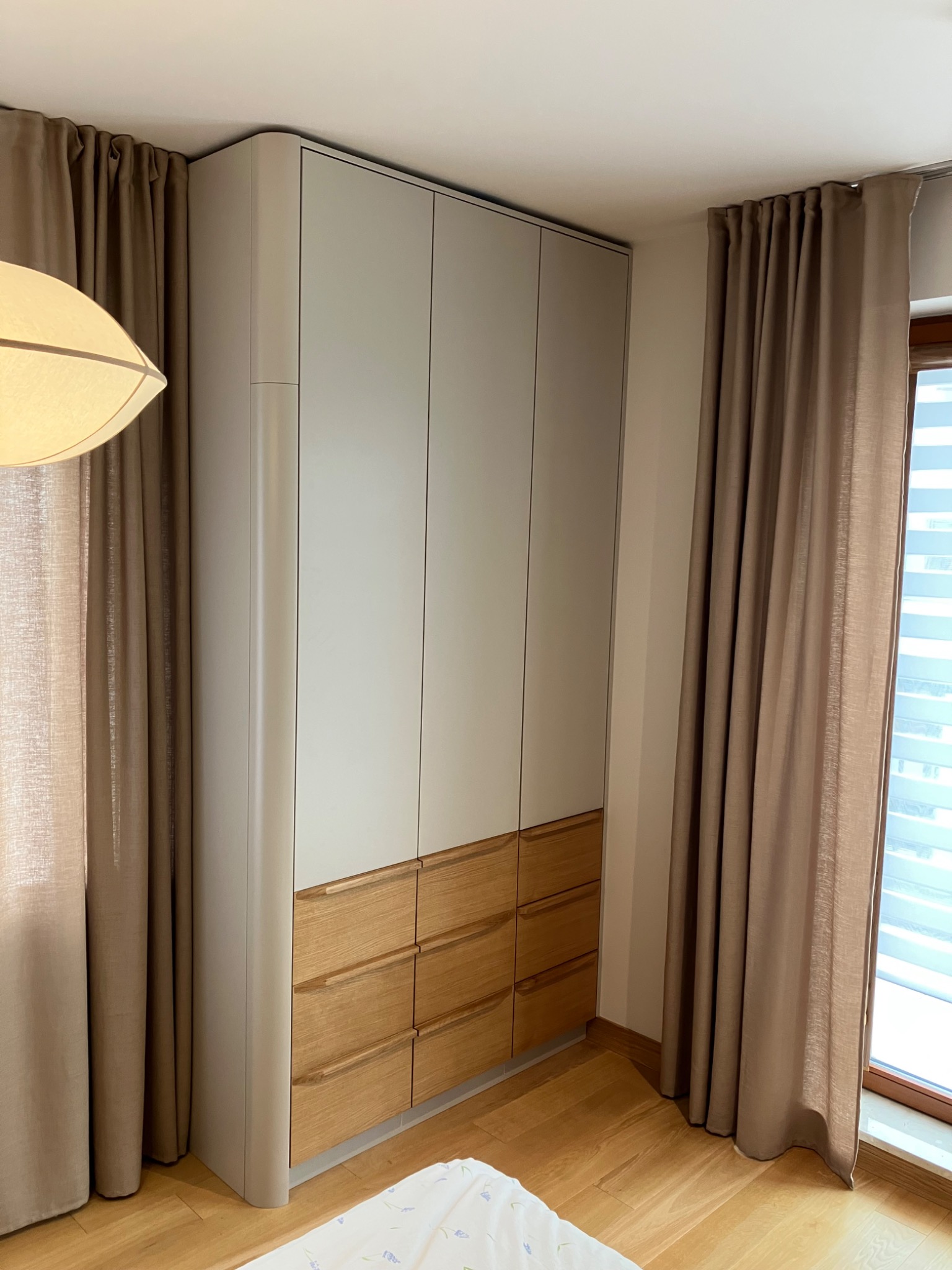 Nowoczesna szafa wnękowa z jasnymi frontami i drewnianymi szufladami, wkomponowana w przestrzeń sypialni z beżowymi zasłonami i drewnianą podłogą.