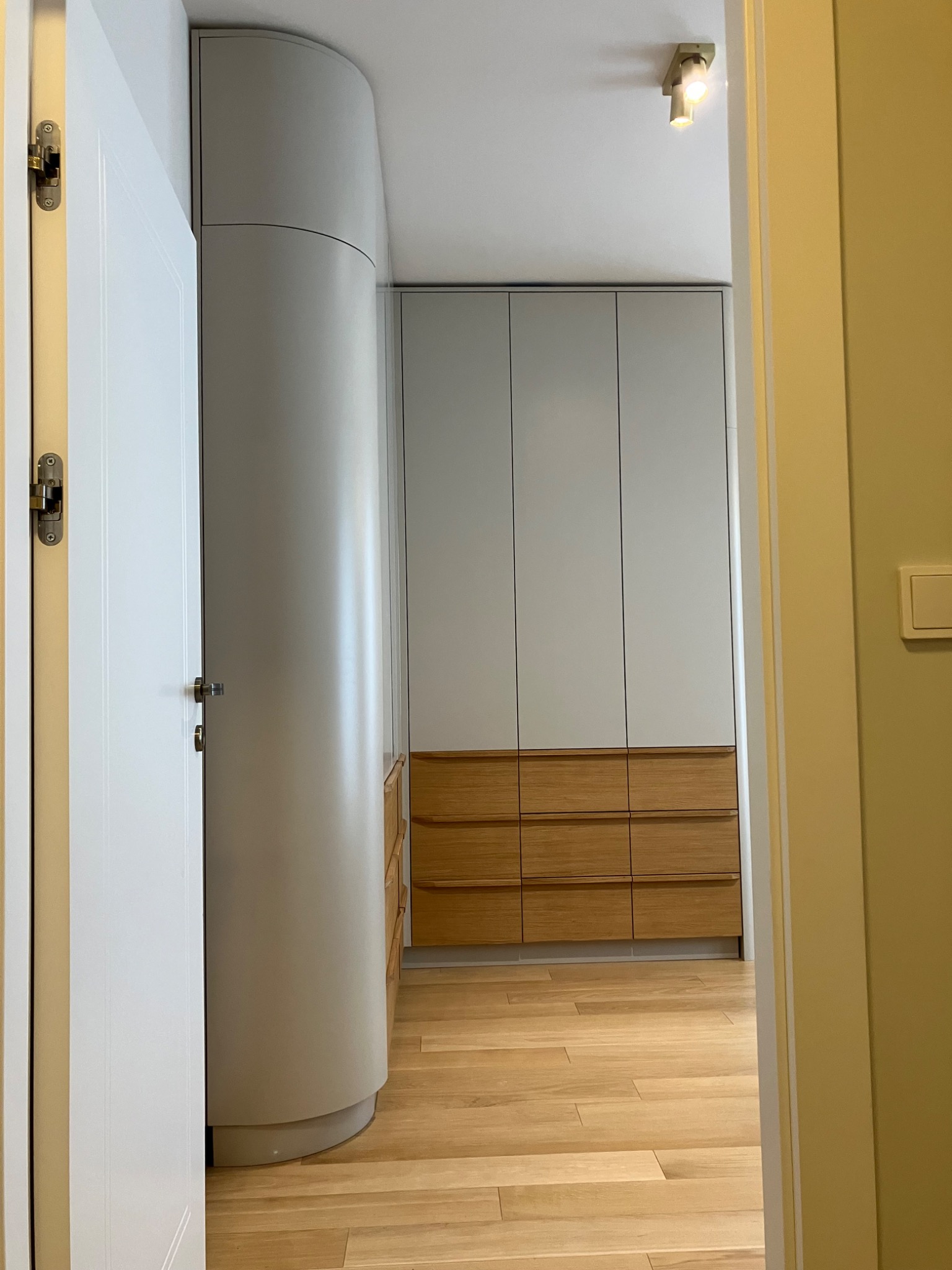 Nowoczesna szafa wnękowa z drewnianymi szufladami, widoczna zza otwartych drzwi. Zaokrąglony bok szafy, jasne kolory, drewniana podłoga. Minimalistyczny design.