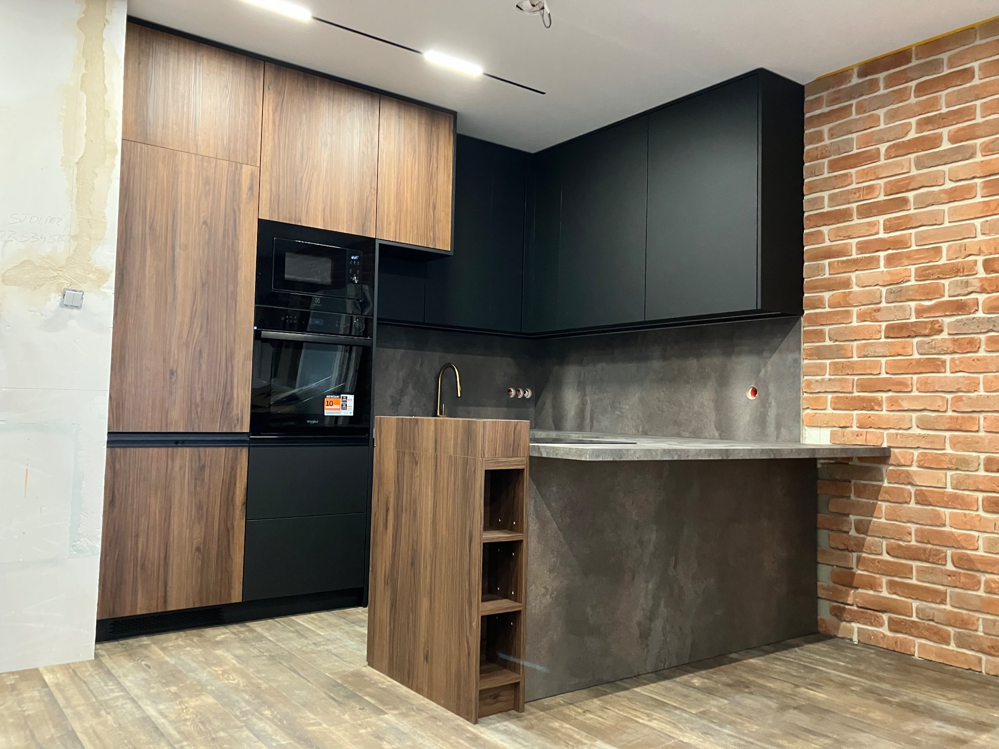 Nowoczesna kuchnia z drewnianymi i czarnymi frontami, szarym blatem oraz ceglaną ścianą. Widoczny fragment ściany w trakcie wykończenia.