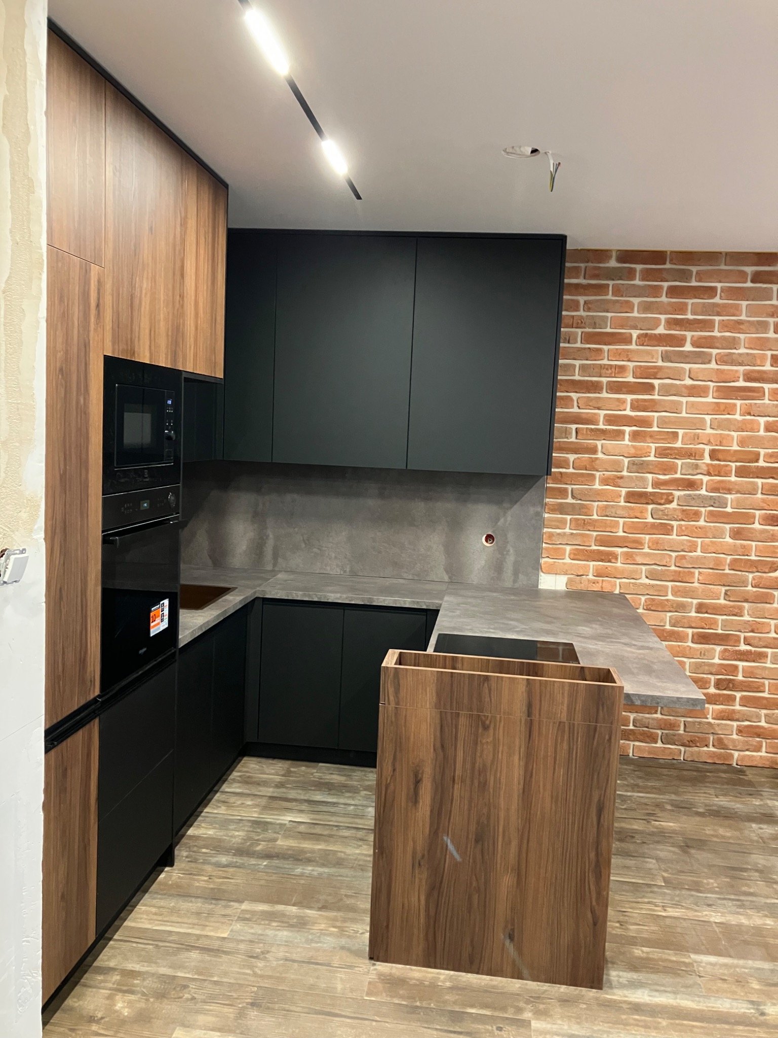 Nowoczesna kuchnia na wymiar: czarne szafki, drewniane akcenty, szary blat i ceglana ściana. Oświetlenie LED. Widoczne niedokończone wykończenia ścian.
