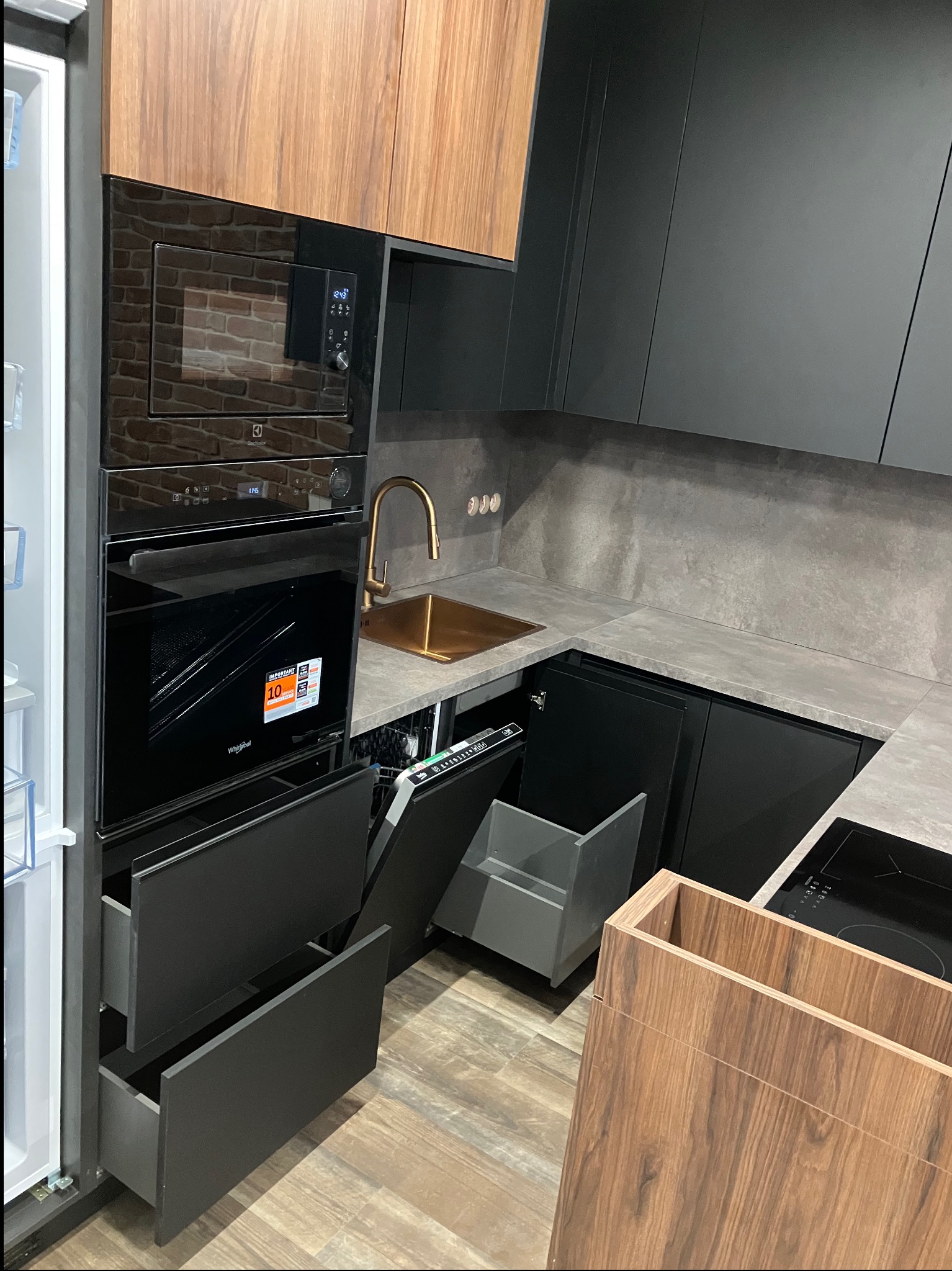 Nowoczesna kuchnia z czarnymi frontami i drewnianymi akcentami. Widoczne otwarte szuflady, złota armatura i sprzęt AGD w zabudowie. Minimalistyczny design.