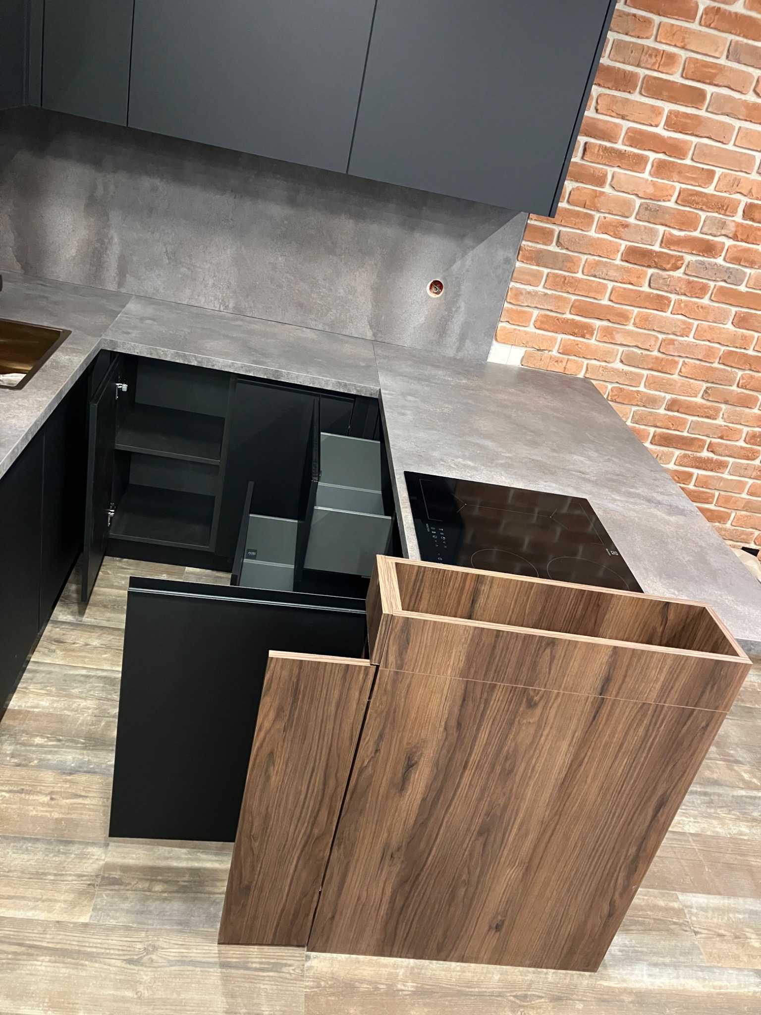 Nowoczesna kuchnia z szarym blatem i czarnymi frontami, otwarte szafki i szuflady z drewnianymi akcentami. Ceglana ściana w tle dodaje charakteru.