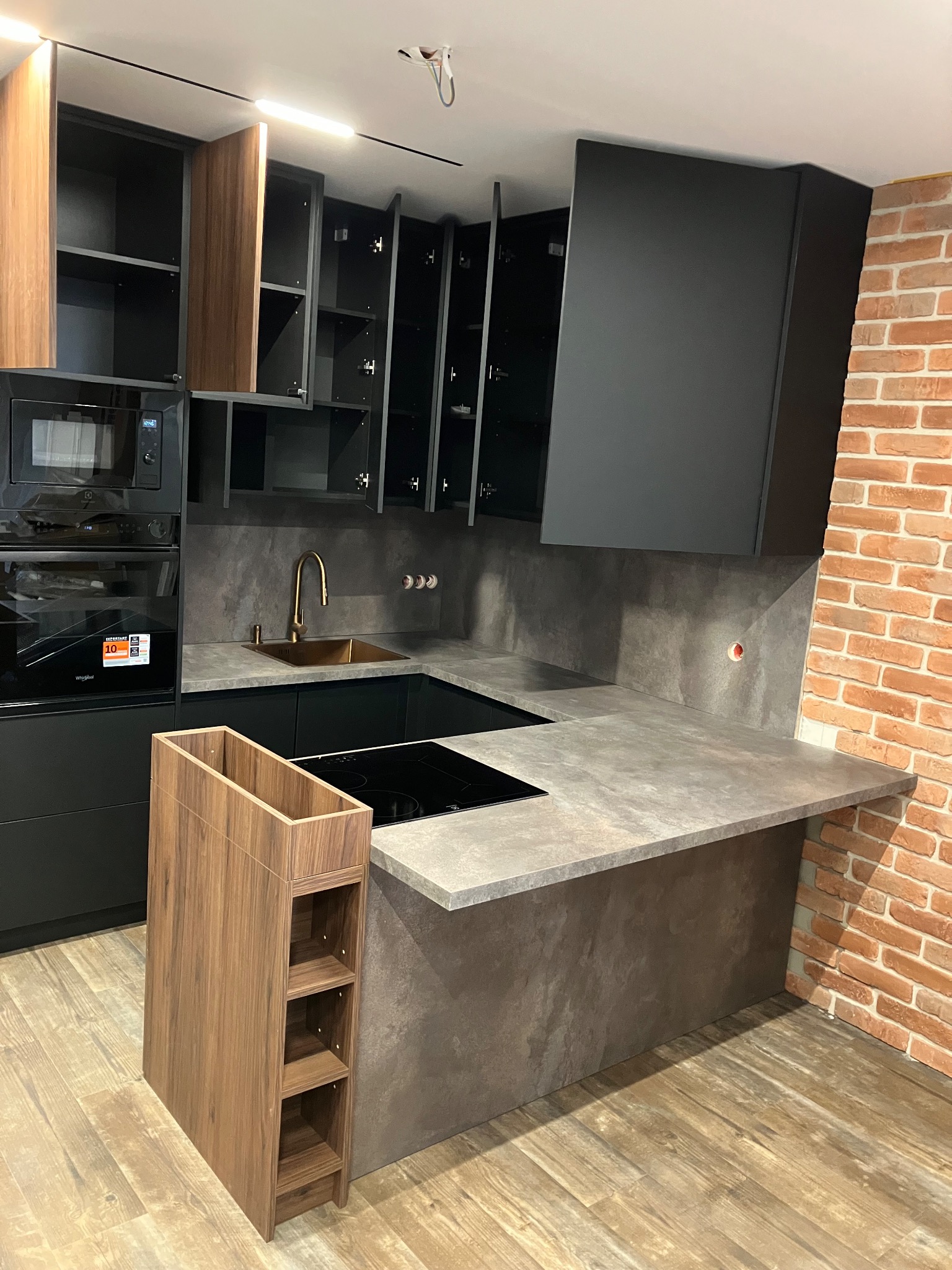 Nowoczesna kuchnia z ciemnymi szafkami, złotą armaturą i blatem w odcieniach szarości. Widoczna ceglana ściana i drewniany regał na wino.
