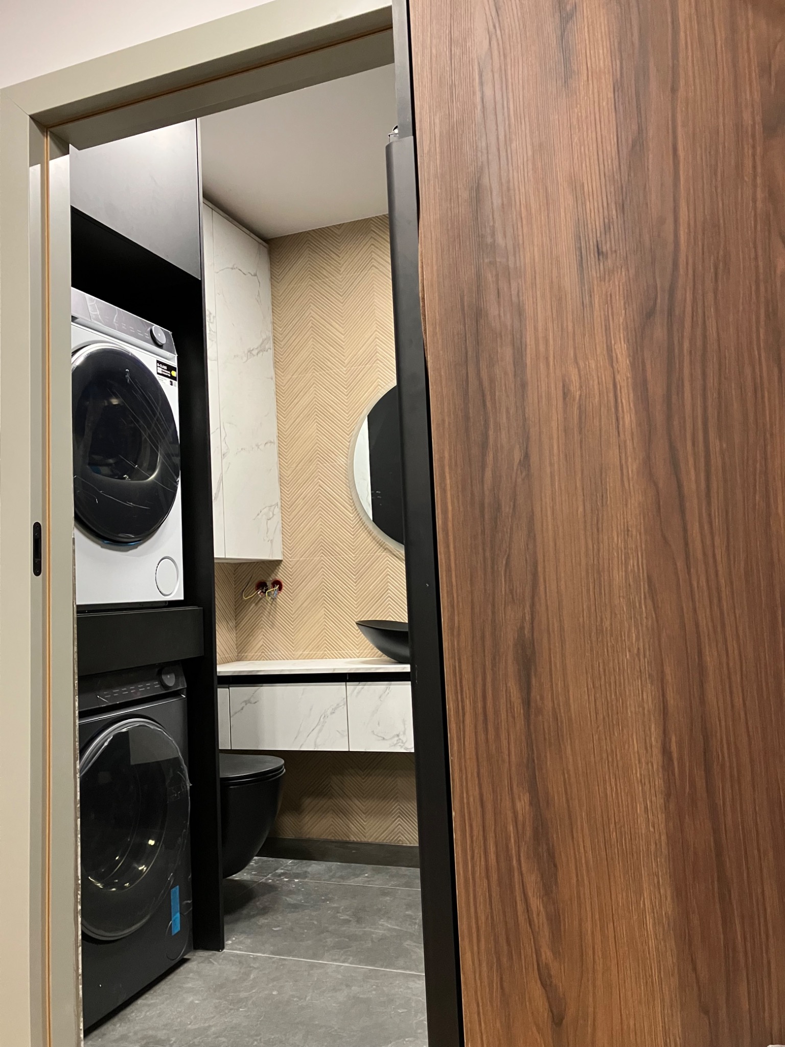 Nowoczesna łazienka z pralką i suszarką w zabudowie za przesuwnymi drzwiami z forniru orzechowego. Minimalistyczny design, marmurowe akcenty i geometryczna tapeta.