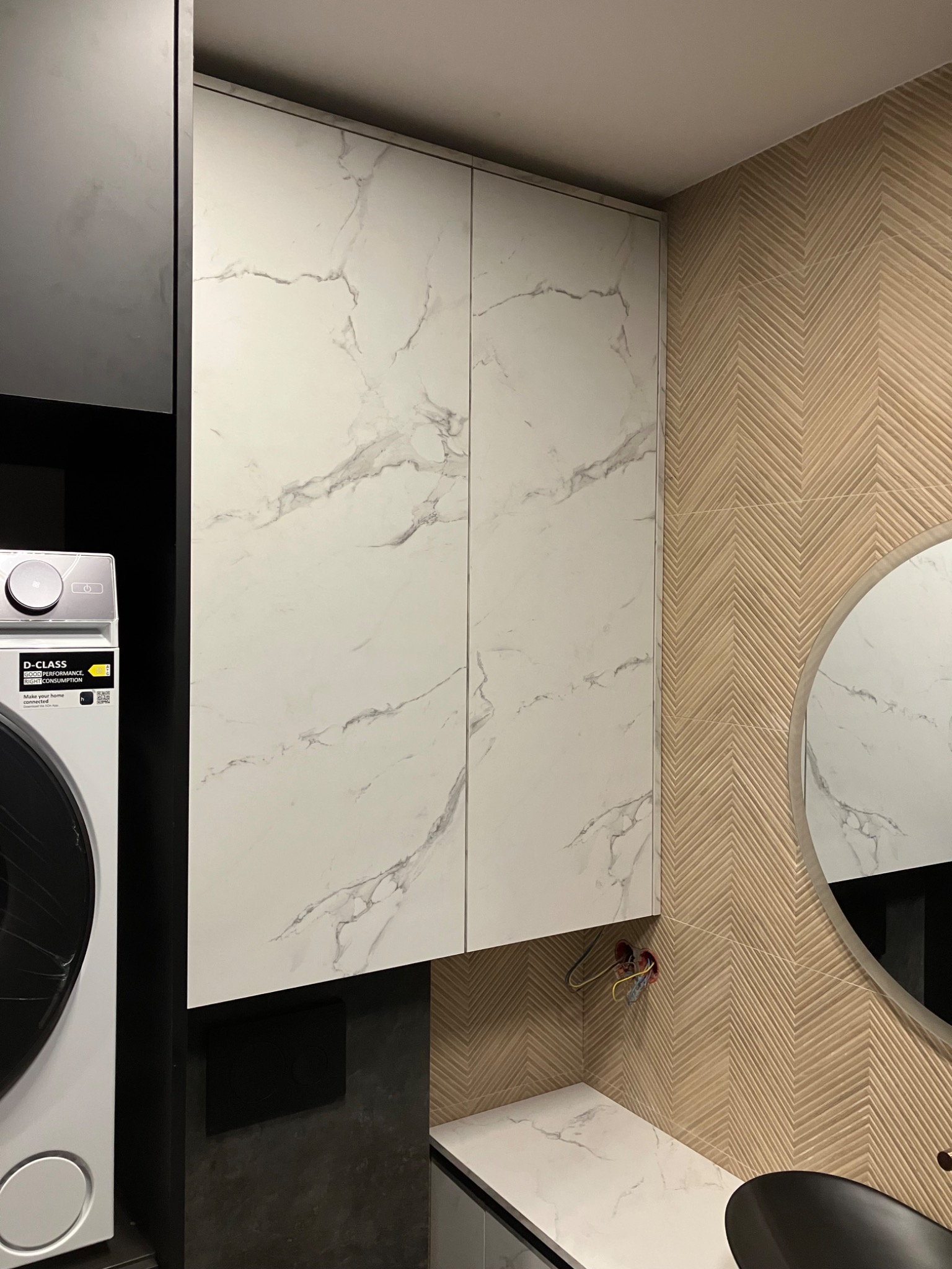 Biała szafka łazienkowa na wymiar z marmurowym wzorem, obok pralka i lustro na ścianie z wzorem jodełki. Nowoczesne wykończenie łazienki.