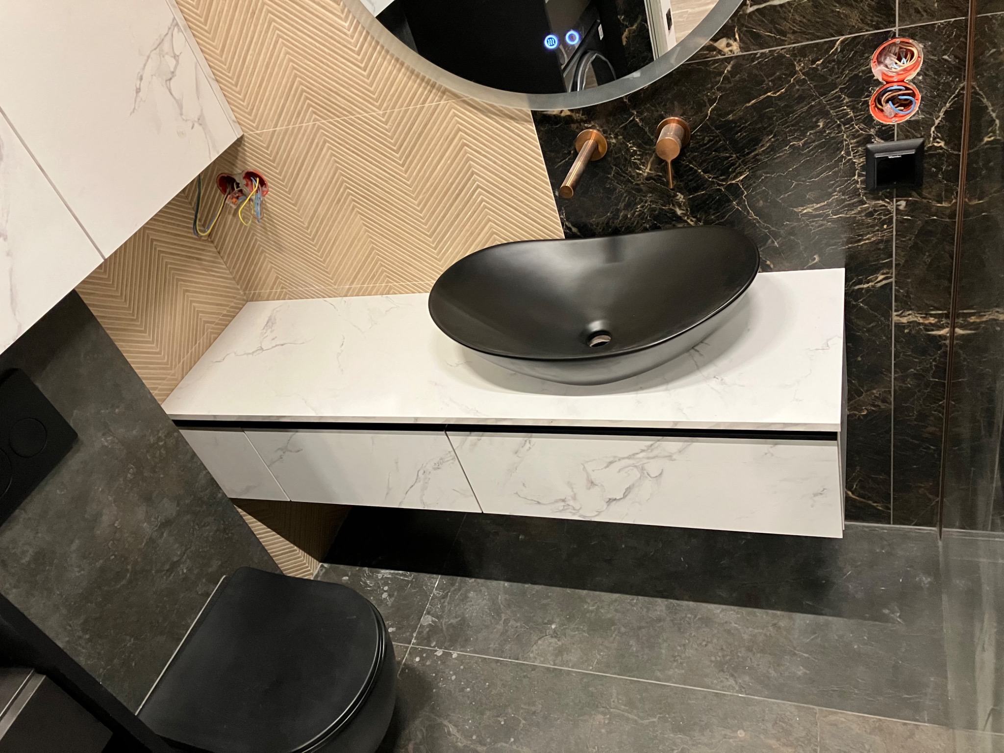 Nowoczesna łazienka z szafką pod umywalkę z blatem imitującym marmur, czarną umywalką i baterią w kolorze miedzi. Na ścianie wzór w jodełkę i marmurowe płytki.