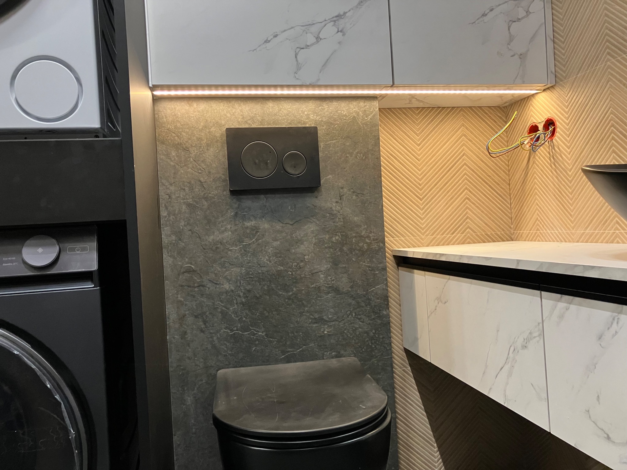 Nowoczesna łazienka z czarną toaletą i umywalką, białe szafki z marmurowym wzorem, podświetlenie LED i wzorzyste płytki. Widoczne gniazdko elektryczne z przewodami.