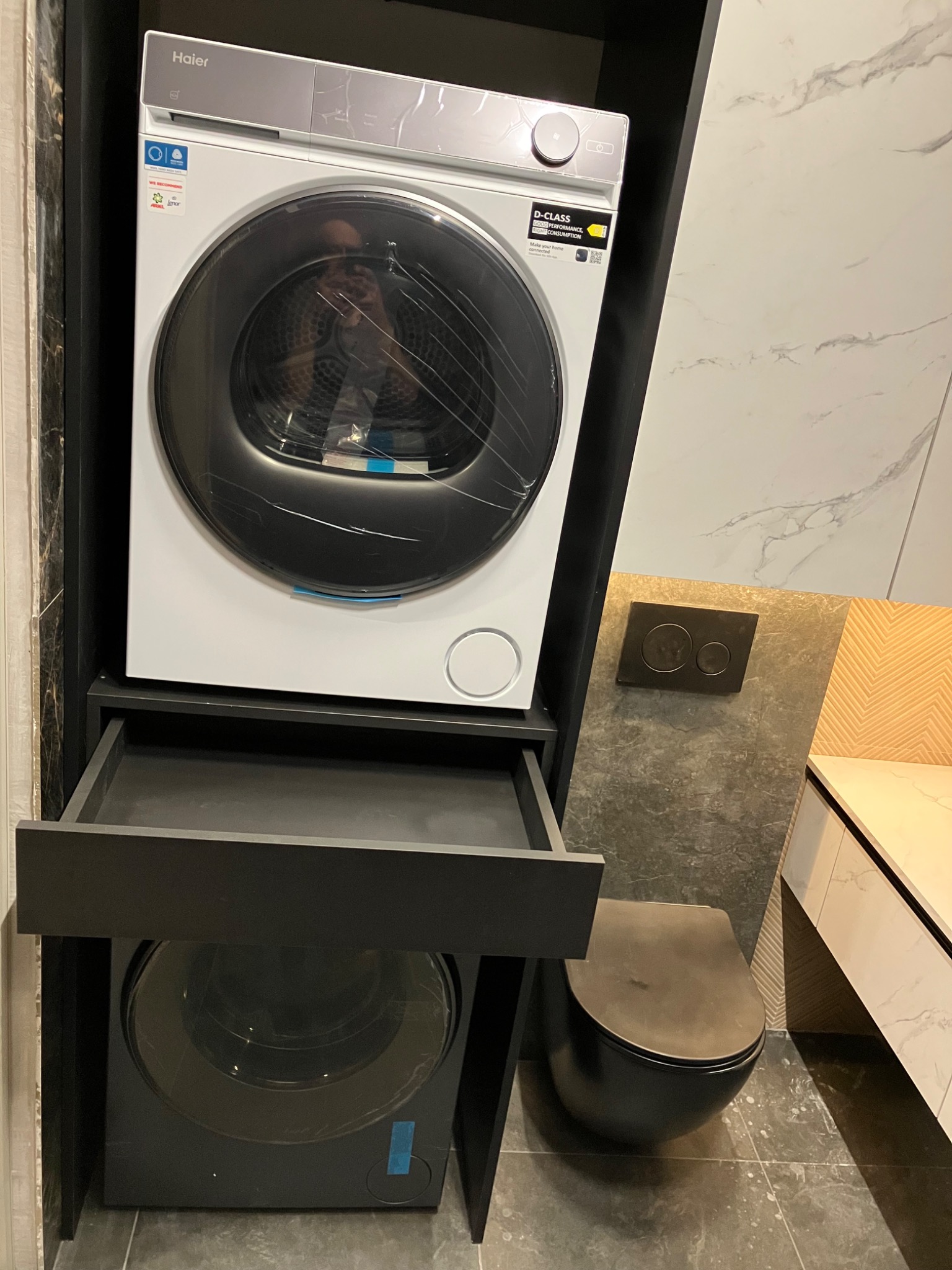 Pralka i suszarka w zabudowie meblowej łazienki, czarne fronty, marmurowe płytki w tle. Nowoczesne rozwiązanie do małych przestrzeni, oszczędność miejsca.