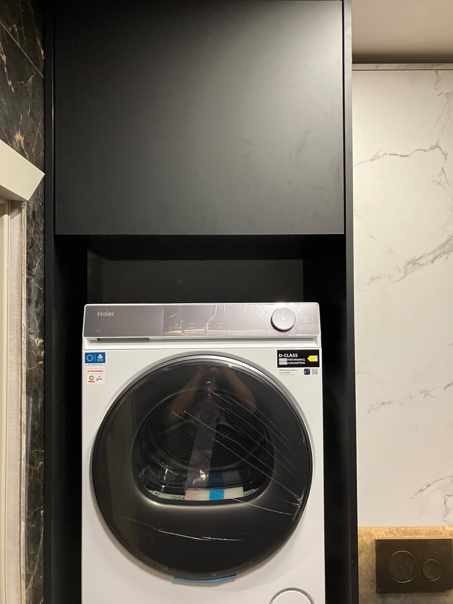 Nowoczesna zabudowa pralki Haier w łazience z czarnymi szafkami i marmurowym wykończeniem. Minimalistyczny design i funkcjonalne rozwiązanie do przechowywania.