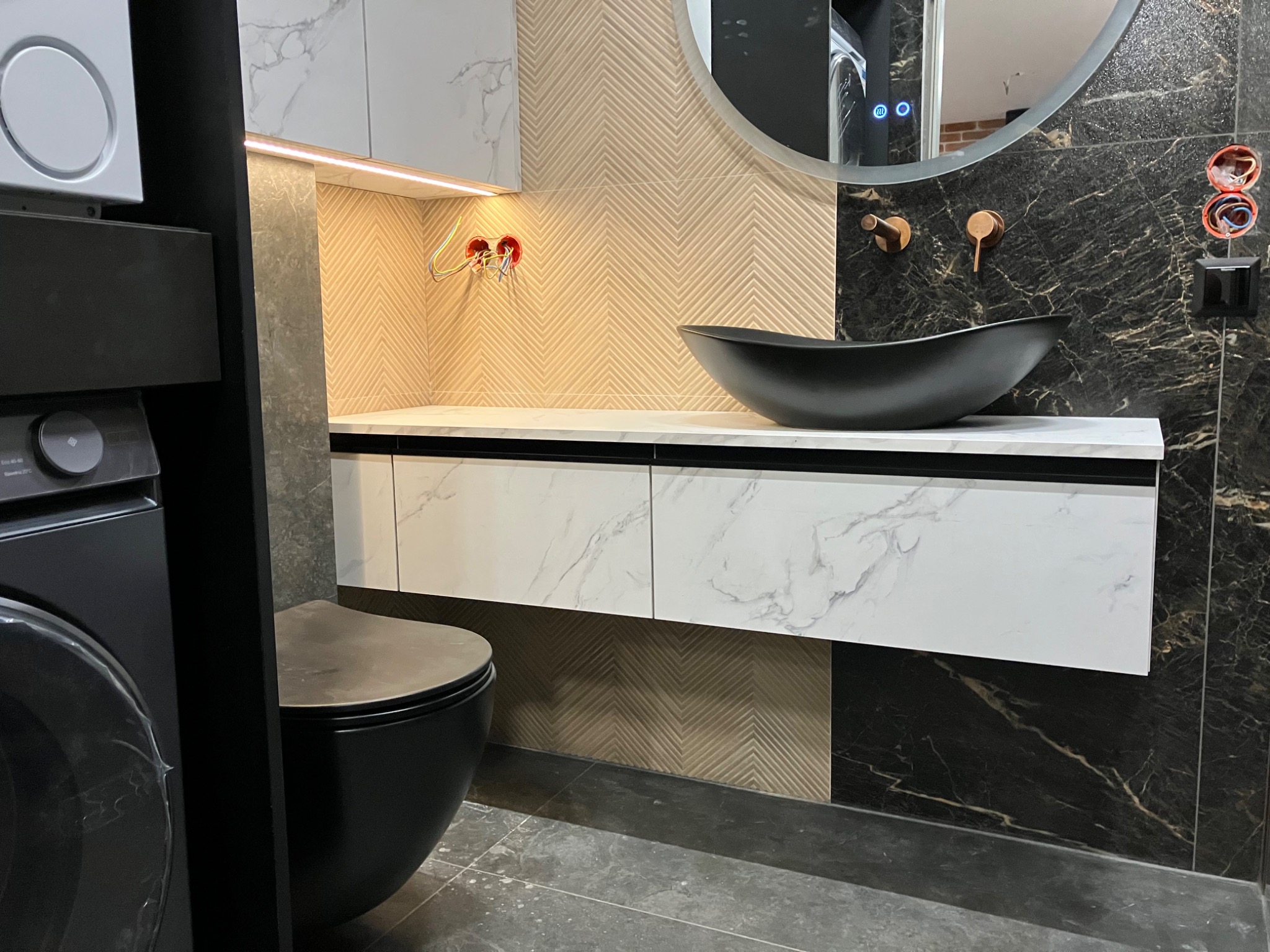 Nowoczesna łazienka z umywalką w kształcie łzy, marmurowym blatem i złotymi akcentami. Widoczne przyłącza elektryczne przed montażem oświetlenia.