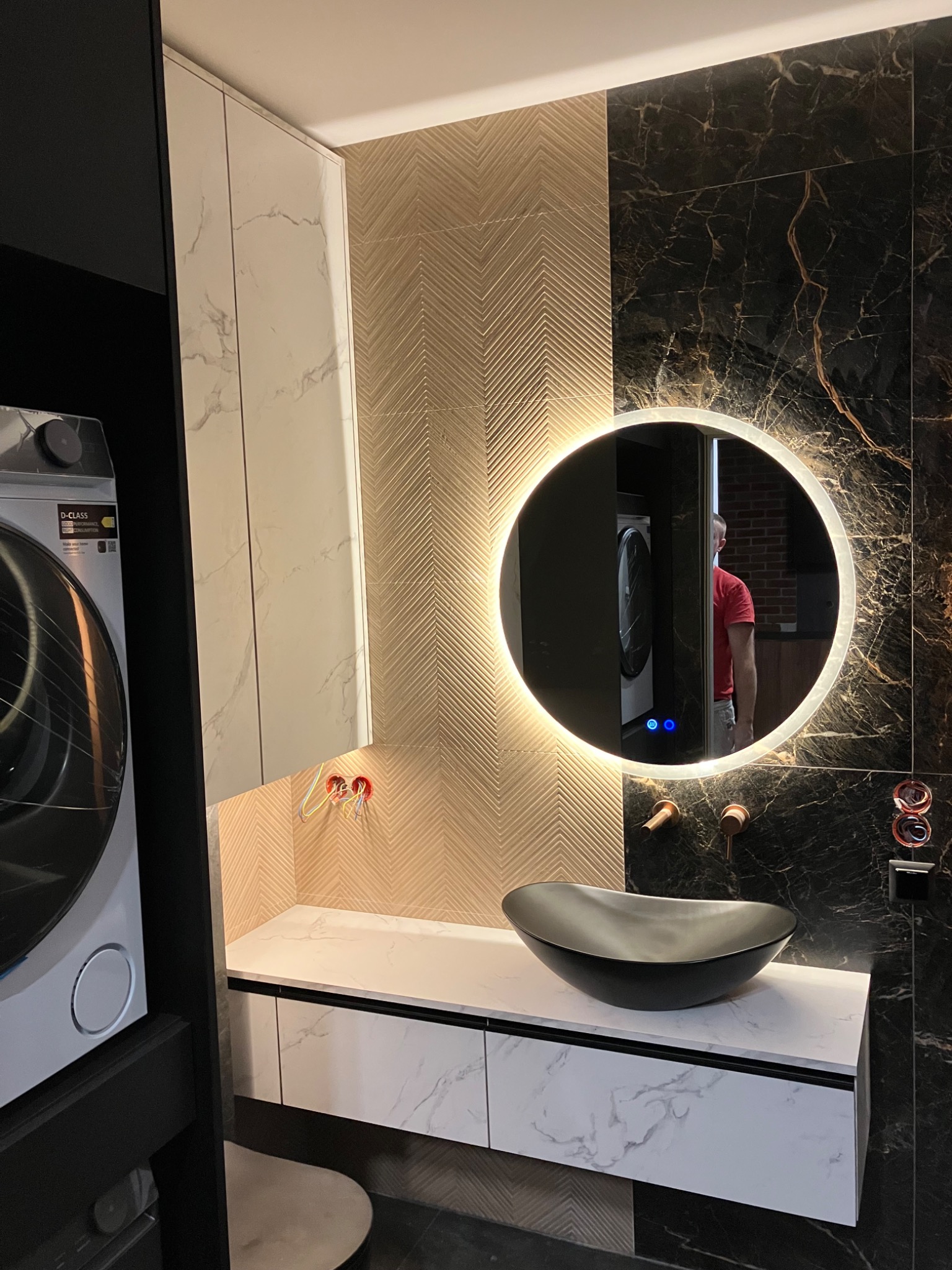 Nowoczesna łazienka z umywalką nablatową, szafką z marmurowym wzorem i okrągłym lustrem z podświetleniem LED. Widoczna pralka w zabudowie i dekoracyjne płytki na ścianie.