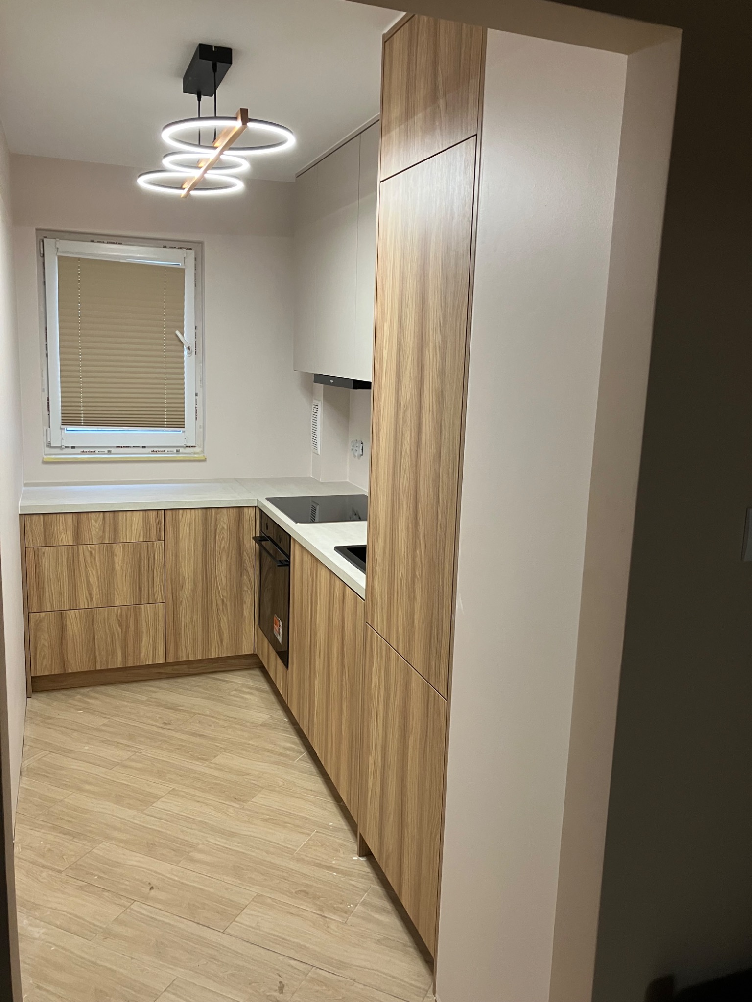 Jasna kuchnia z drewnianymi frontami, oświetlona nowoczesną lampą. Widoczne okno z żaluzjami i płyta indukcyjna. Ukończona realizacja z dbałością o detale.