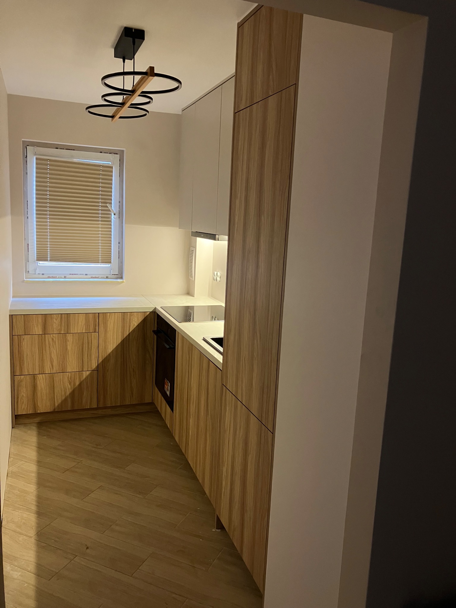 Nowoczesna kuchnia z drewnianymi frontami szafek, białym blatem i designerską lampą sufitową. Ujęcie z perspektywy podłogi, podkreślające układ i wykończenie.