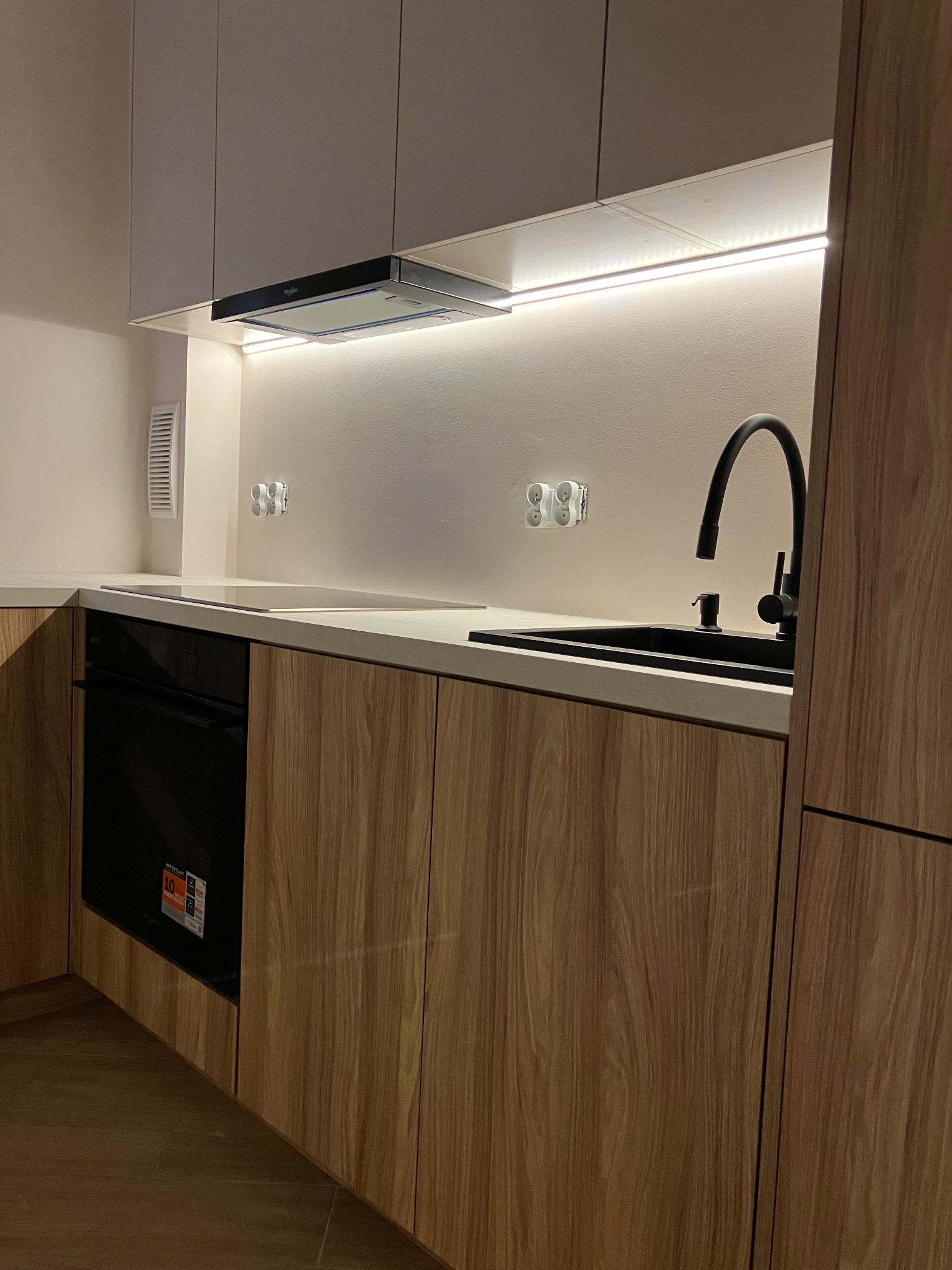 Nowoczesna kuchnia z drewnianymi frontami, czarną armaturą i oświetleniem LED pod szafkami. Minimalistyczny design z ukrytymi gniazdkami i płytą indukcyjną.