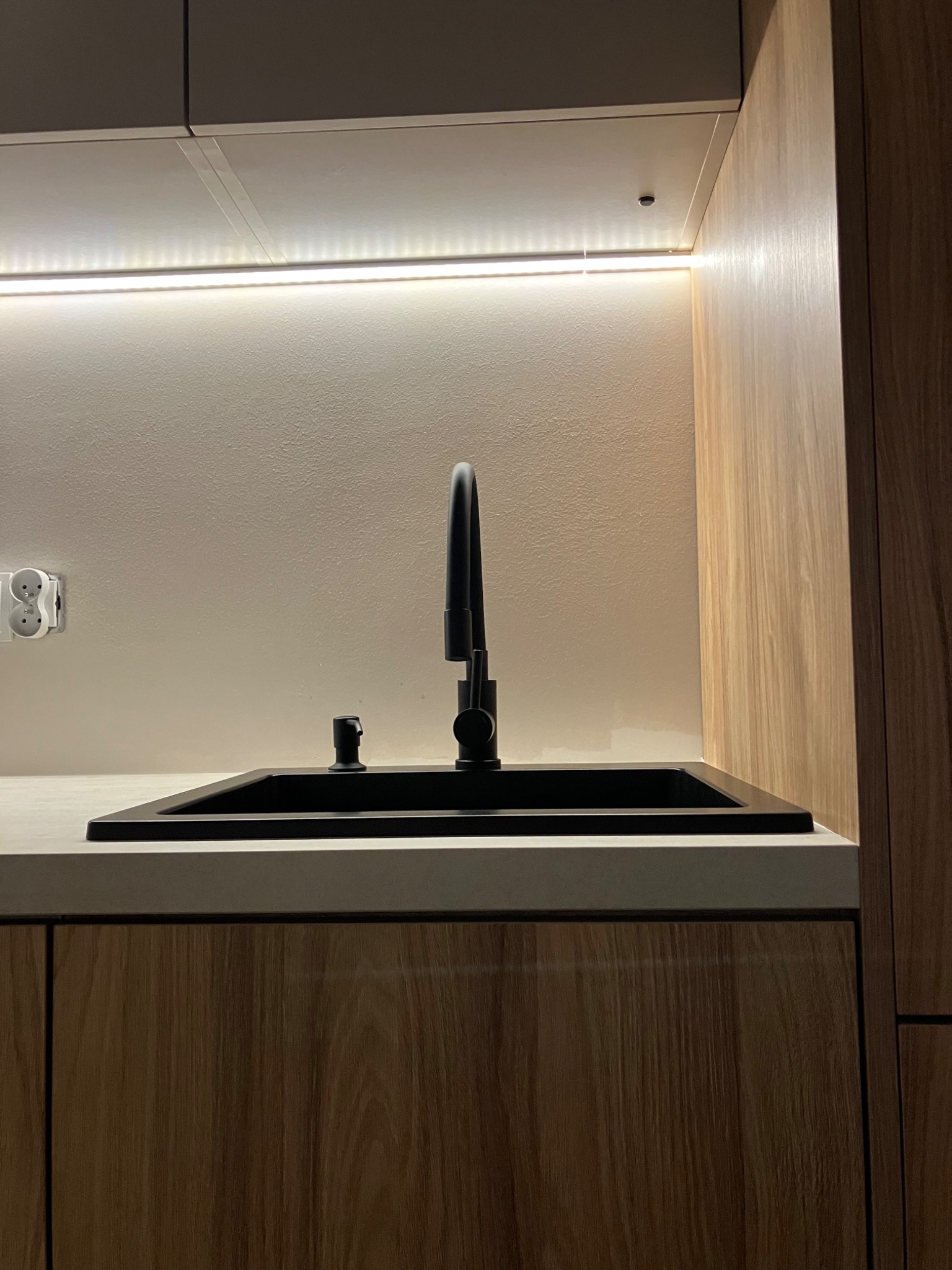 Nowoczesna, minimalistyczna kuchnia: czarny zlew z baterią, drewniane fronty szafek, podświetlenie LED. Widoczny fragment blatu i gniazdko elektryczne.