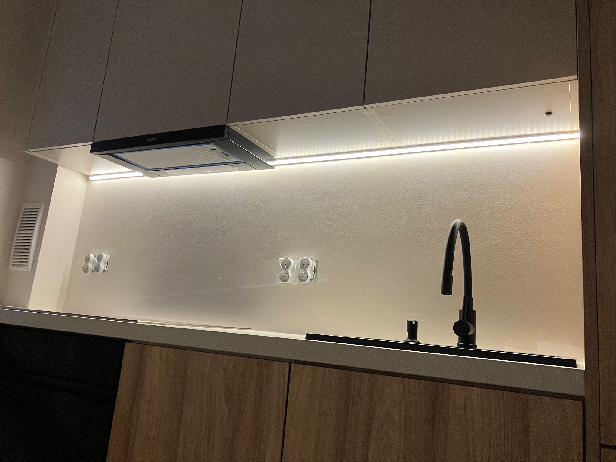 Nowoczesna kuchnia z drewnianymi frontami, czarną armaturą i oświetleniem LED pod szafkami. Widoczne gniazdka elektryczne i okap kuchenny Whirlpool.