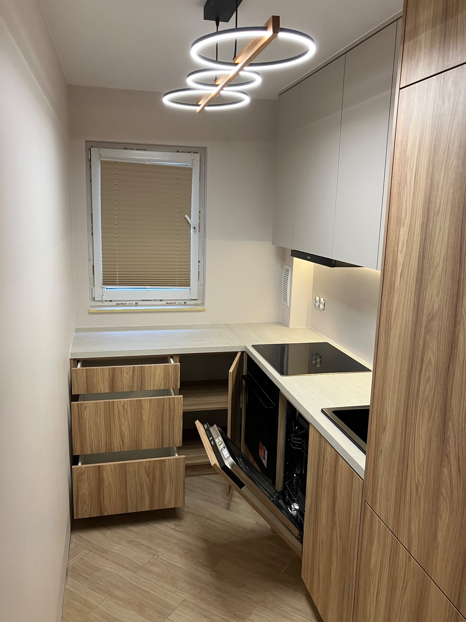 Nowoczesna kuchnia z drewnianymi frontami, otwartymi szufladami i piekarnikiem. Minimalistyczne oświetlenie sufitowe. Jasne wnętrze z blatem pod oknem.