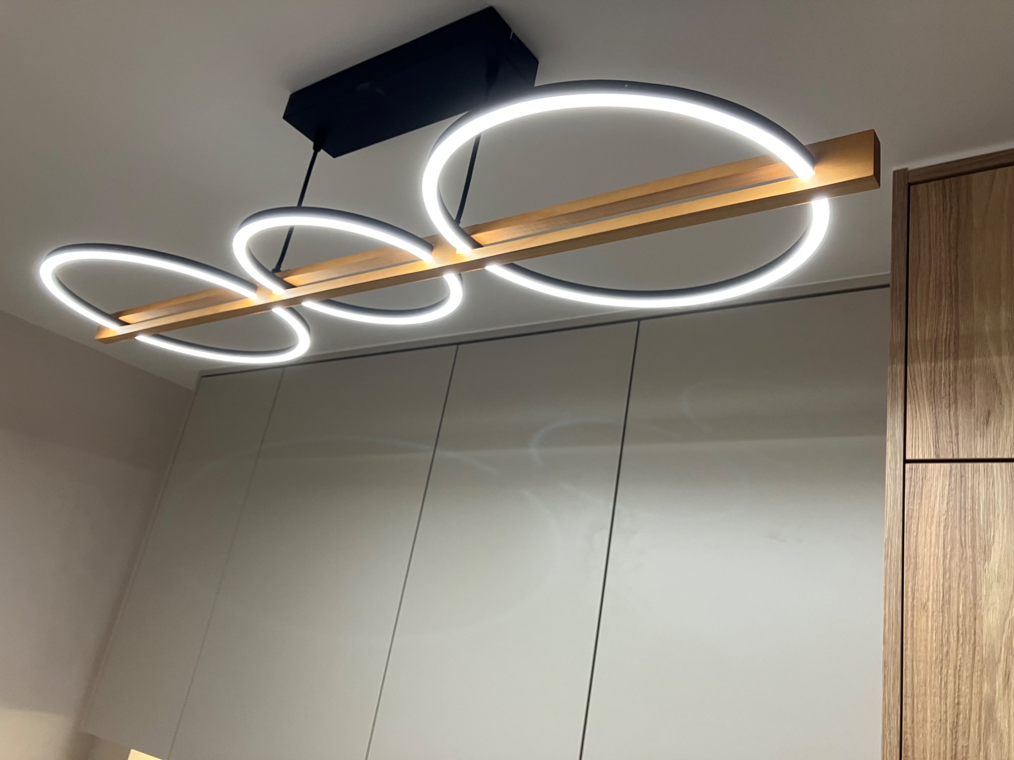 Nowoczesna lampa sufitowa LED z drewnianym elementem, oświetlająca jasne, minimalistyczne szafki kuchenne. Unikalny design i ciepłe światło.