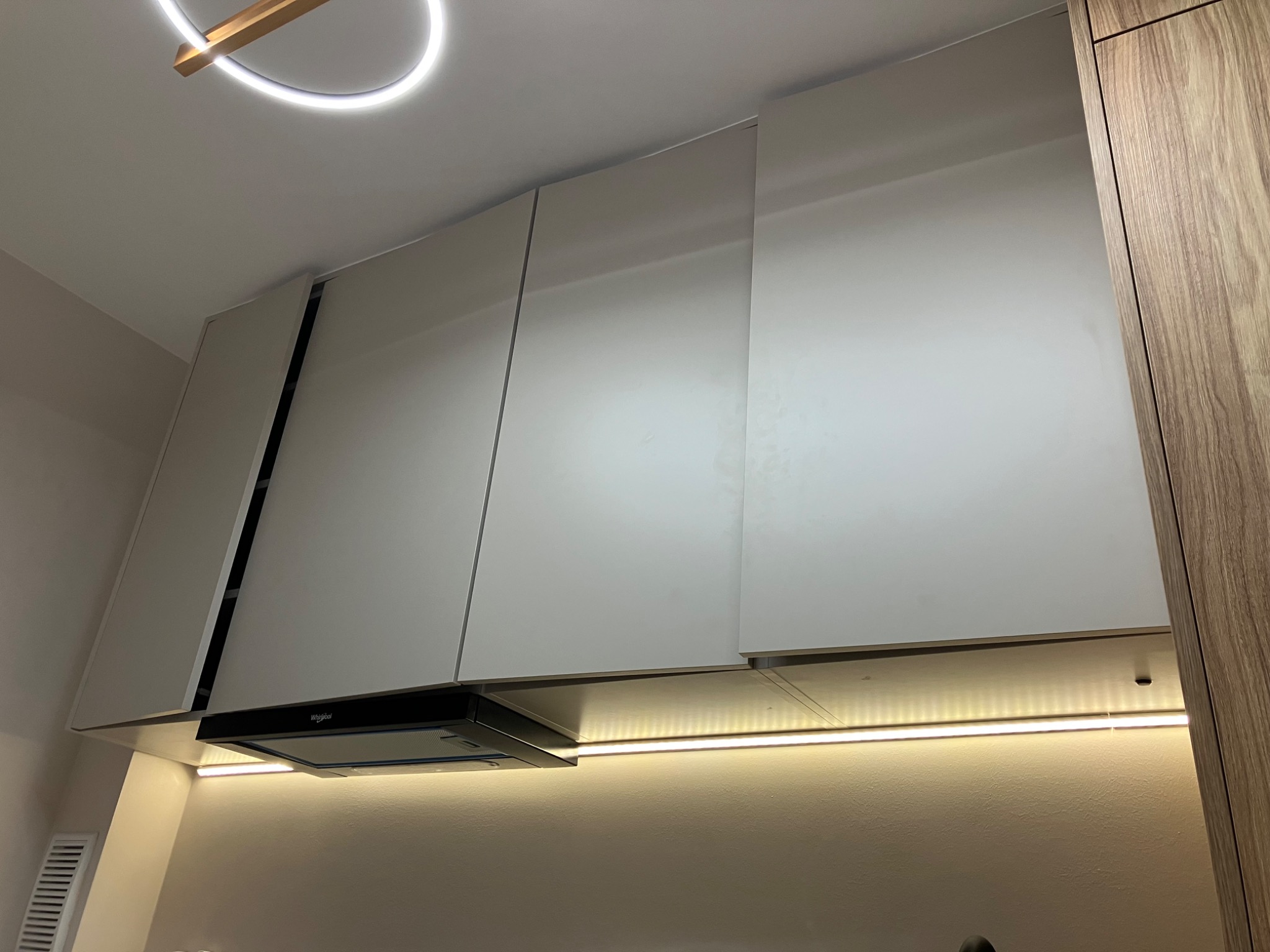 Nowoczesna kuchnia z białymi szafkami wiszącymi i okapem Whirlpool, oświetlona listwą LED pod szafkami. U góry widoczny fragment okrągłej lampy sufitowej.