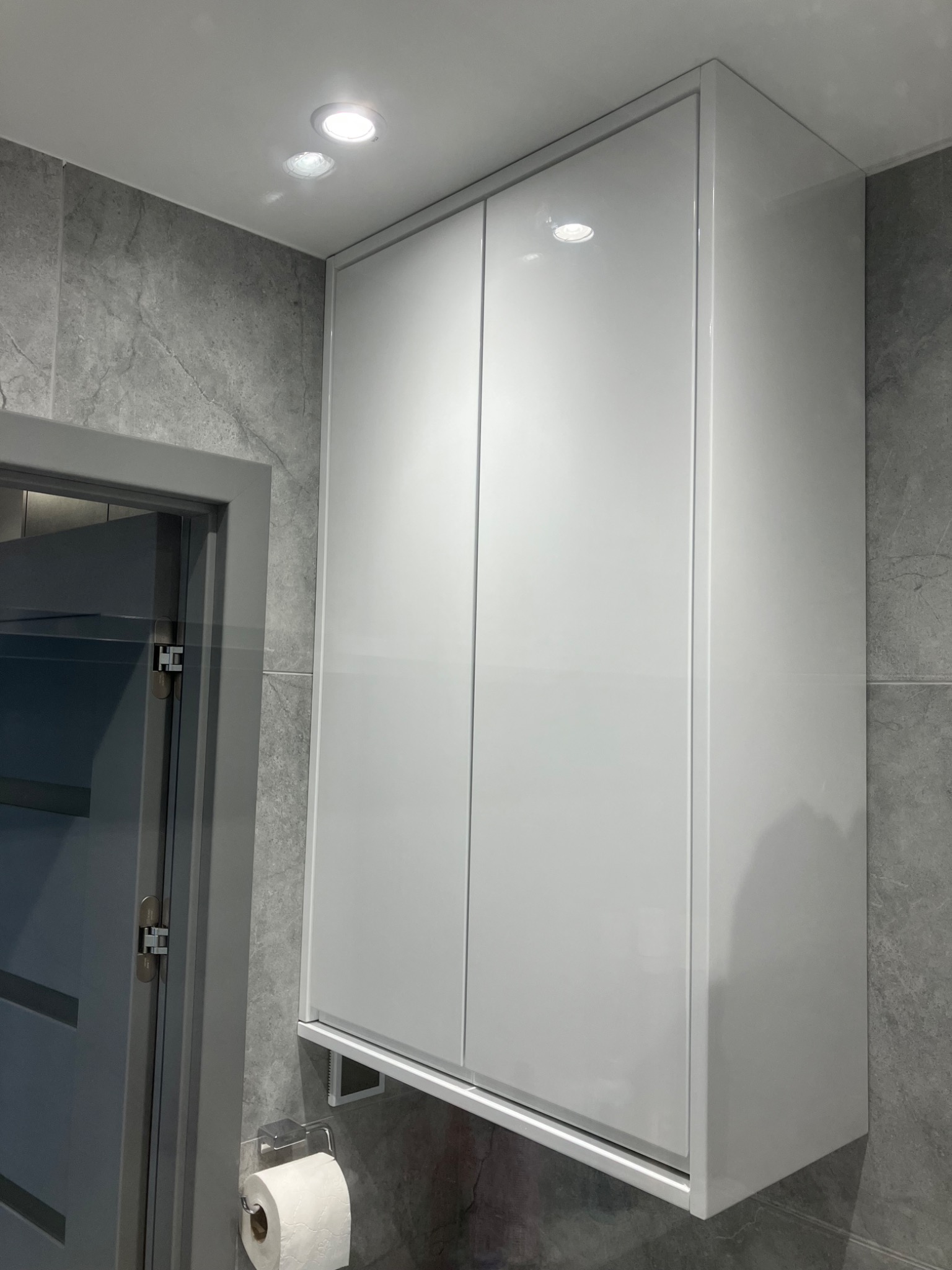 Biała, lakierowana szafka łazienkowa na ścianie z szarych płytek, z widocznym uchwytem na papier toaletowy i fragmentem szarej ościeżnicy drzwiowej.