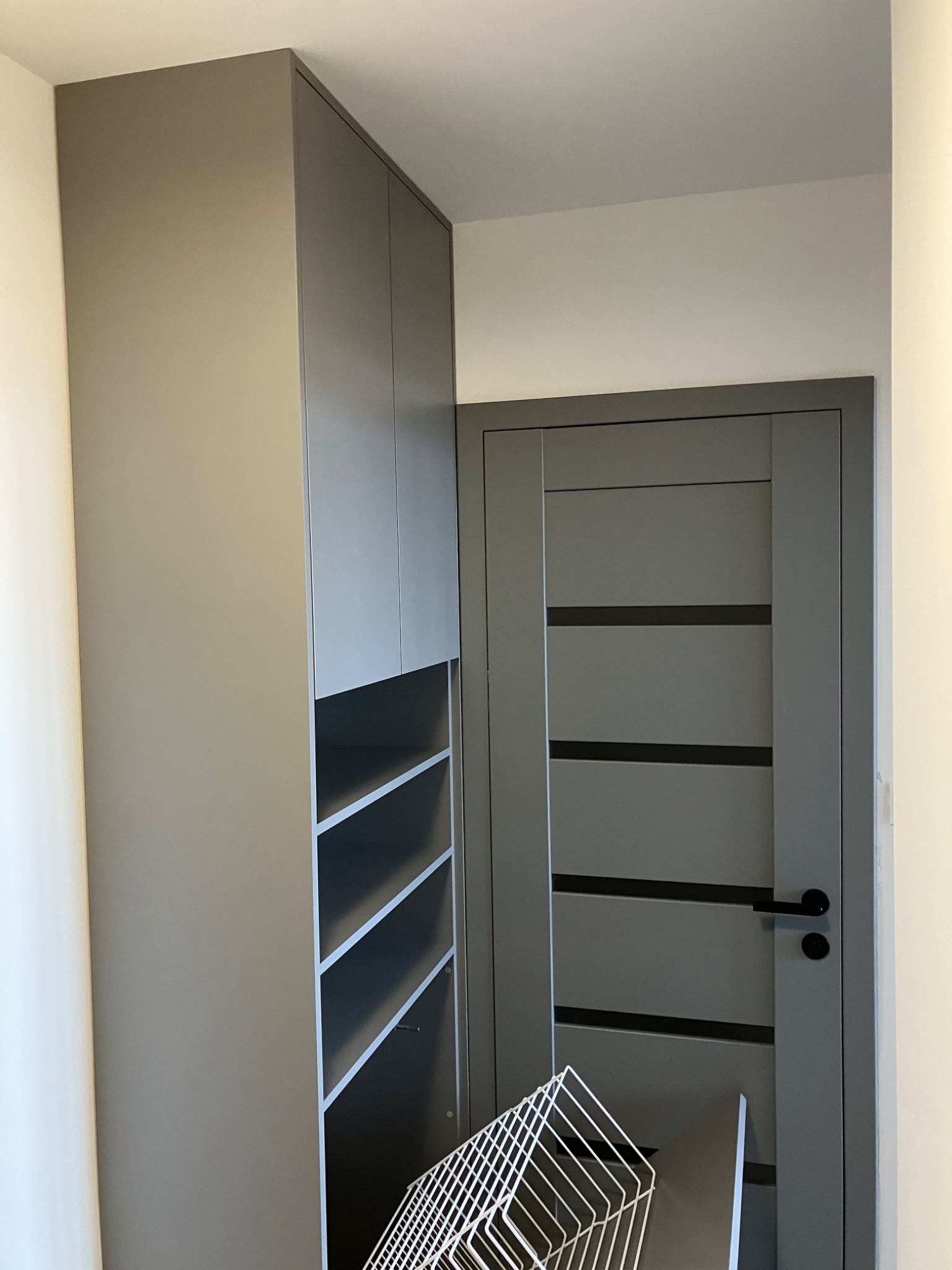 Szara szafa z otwartymi półkami obok drzwi w tym samym odcieniu szarości. W dolnej części szafy widoczny biały kosz.