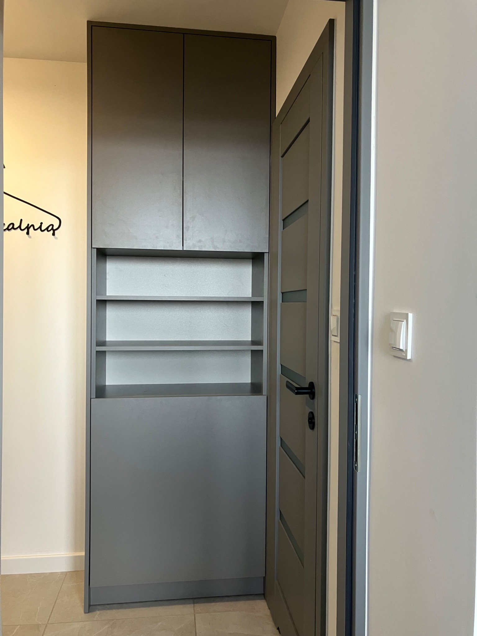 Szara szafa wnękowa z otwartymi półkami, dopasowana kolorystycznie do drzwi wejściowych, w minimalistycznym wnętrzu.