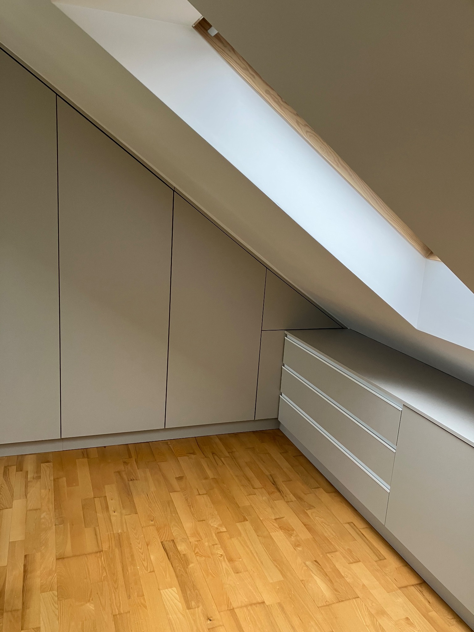 Szafa wnękowa ze szufladami w kolorze beżowym, dopasowana do skosu poddasza, z drewnianą podłogą i oknem dachowym. Minimalistyczny design.