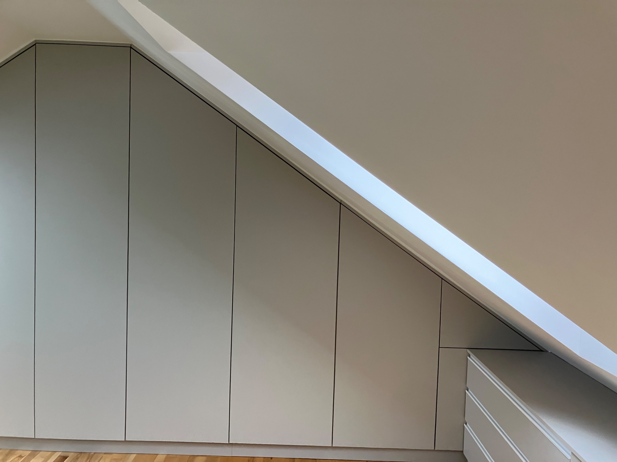 Jasnoszara zabudowa szafy wnękowej na poddaszu, dopasowana do skosu dachu. Minimalistyczny design, widoczne szuflady i drewniana podłoga.