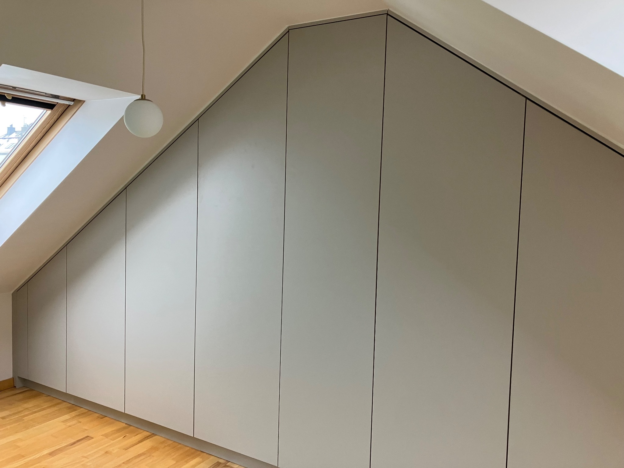 Jasnoszara szafa wnękowa na poddaszu, dopasowana do skosu dachu. Widoczne drewniane panele podłogowe i okno dachowe. Minimalistyczny design.