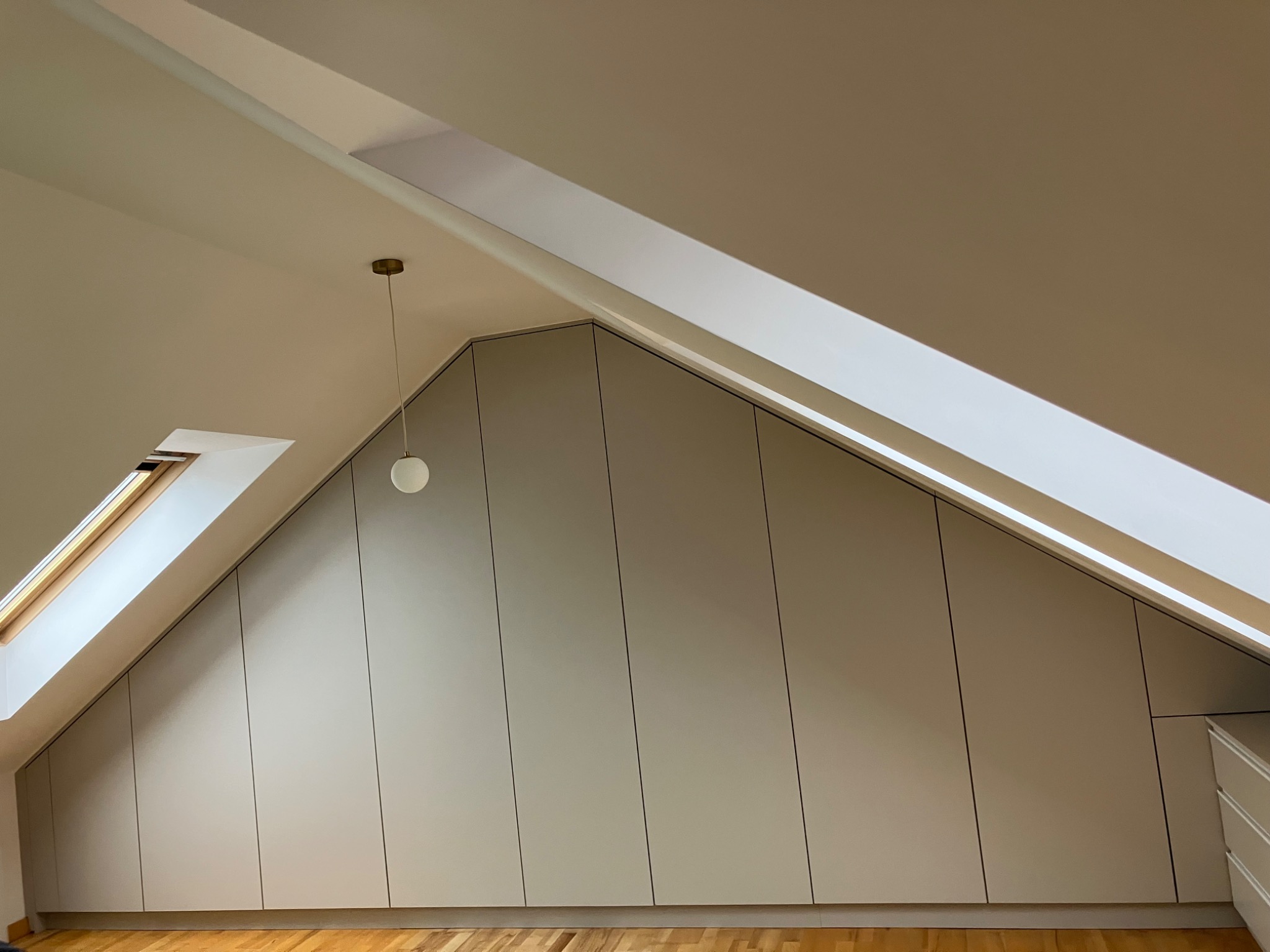 Szafa wnękowa na poddaszu, dopasowana do skosu dachu. Jasne, minimalistyczne fronty, drewniana podłoga, okno dachowe i lampa wisząca.