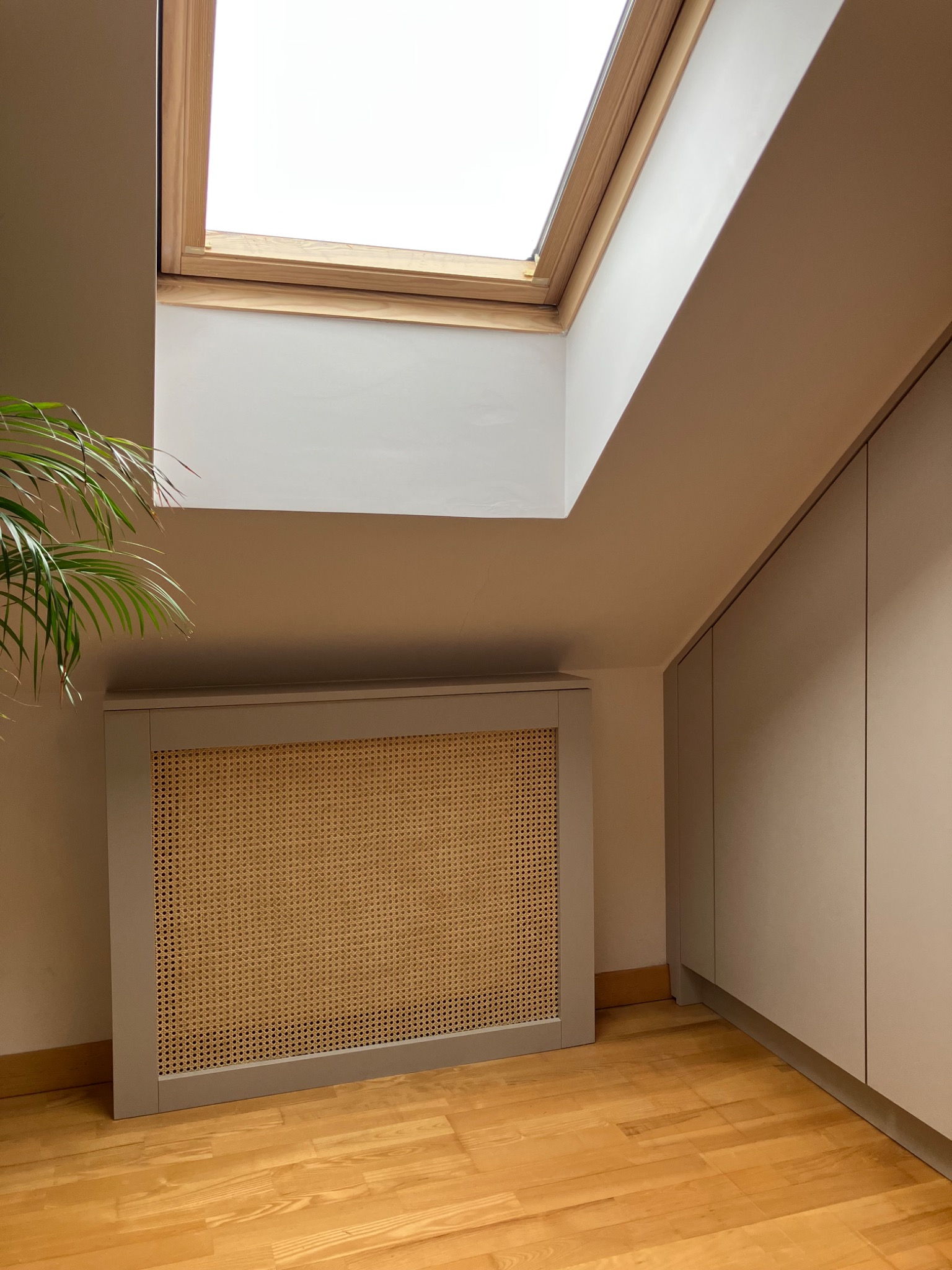 Beżowa szafa wnękowa z rattanowym frontem, drewniana podłoga, okno dachowe. Minimalistyczny design, neutralna kolorystyka, roślina w lewym rogu kadru.