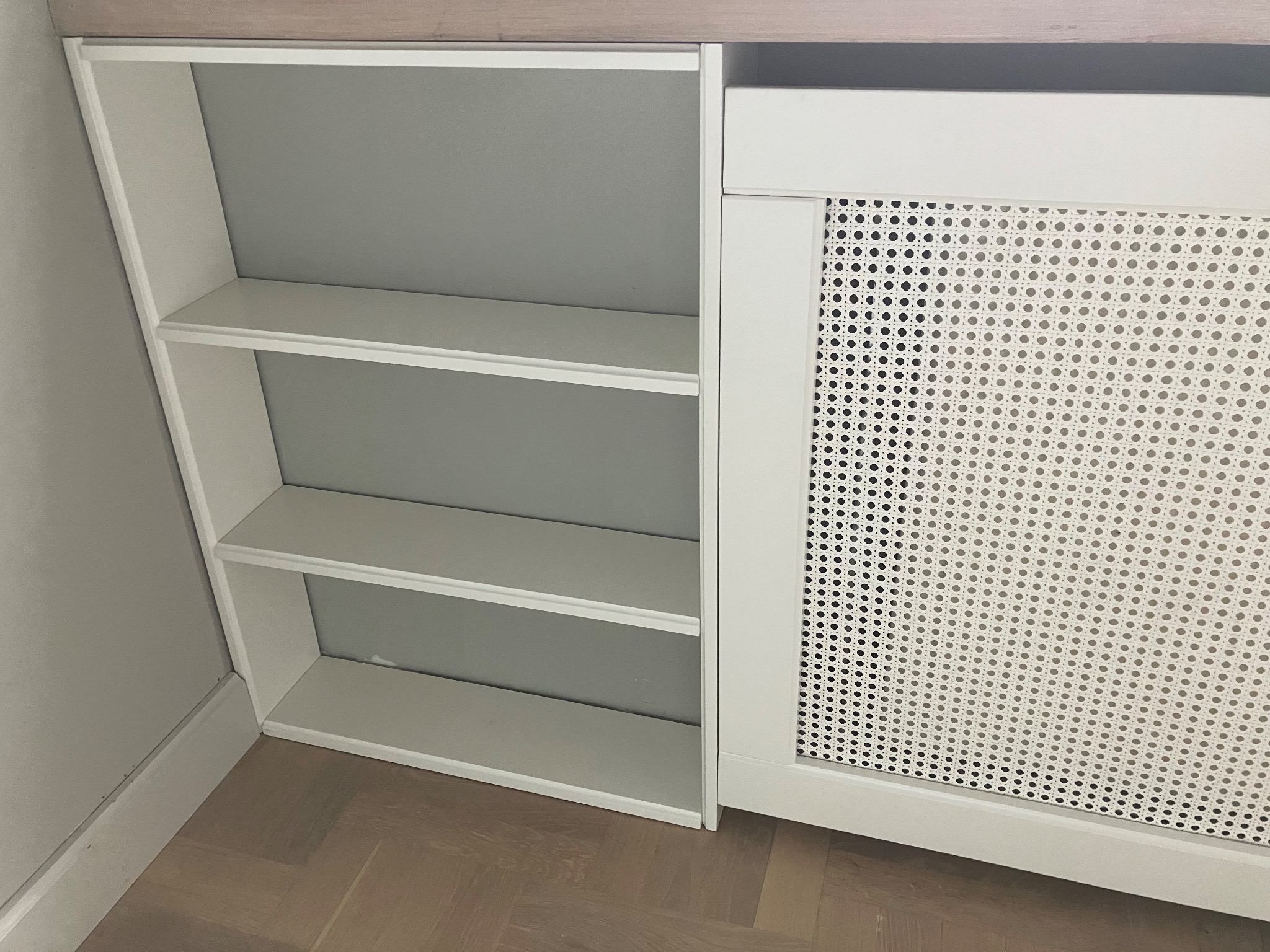 Biała zabudowa grzejnika z ażurowym frontem i otwartymi półkami, wykonana na wymiar w Warszawie. Minimalistyczny design, widoczny parkiet na podłodze.