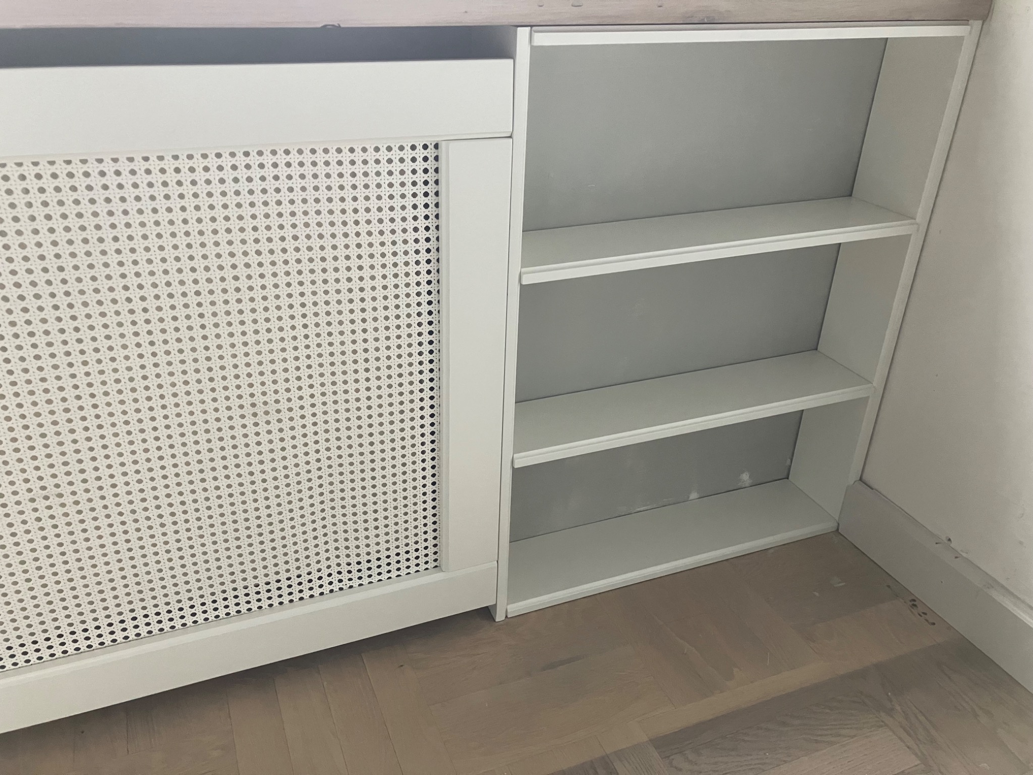 Biała szafka z ażurowymi drzwiczkami i otwartymi półkami, widok z bliska na drewnianej podłodze. Minimalistyczny design, jasne wnętrze półek.