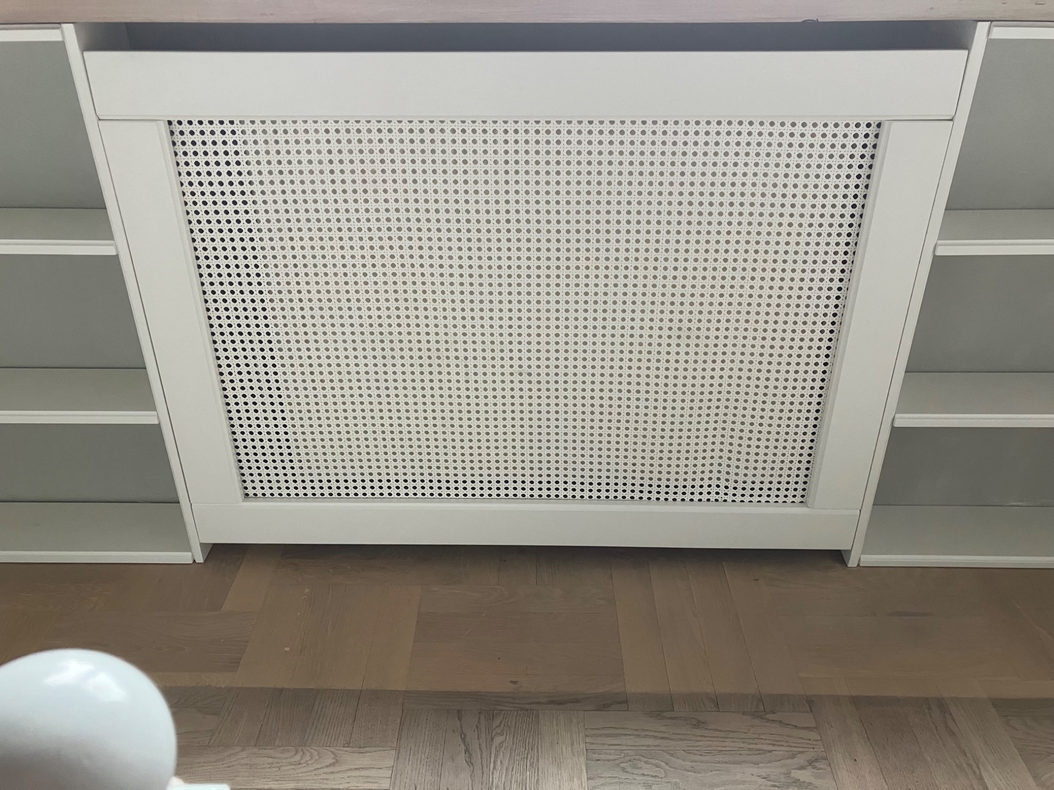 Biała zabudowa grzejnika z ażurowym frontem i półkami po bokach, na drewnianej podłodze. Minimalistyczny design, widok z dołu.
