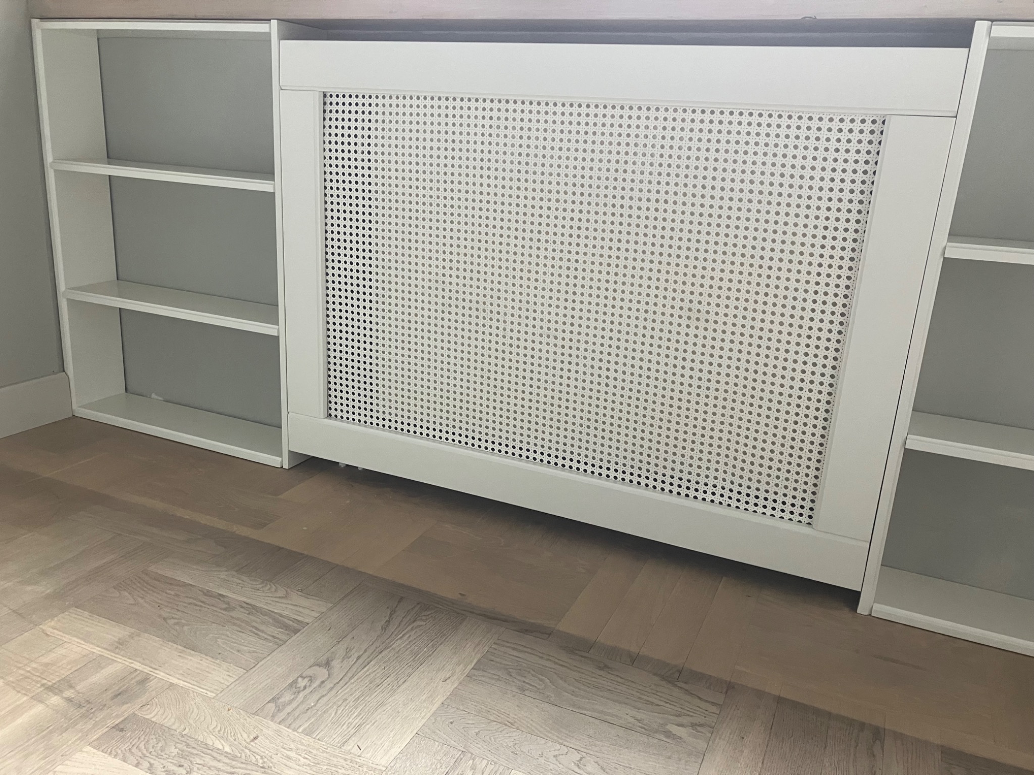 Biała zabudowa grzejnika z ażurowym frontem, wkomponowana między dwie otwarte szafki z półkami. Parkiet w jodełkę na podłodze. Minimalistyczny design.