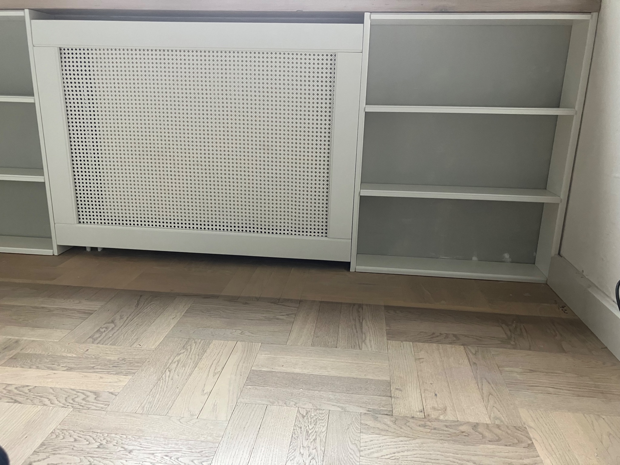 Biała zabudowa meblowa z ażurowym frontem maskującym grzejnik i otwartymi półkami, na parkiecie ułożonym w jodełkę. Minimalistyczny design, jasne wnętrze.