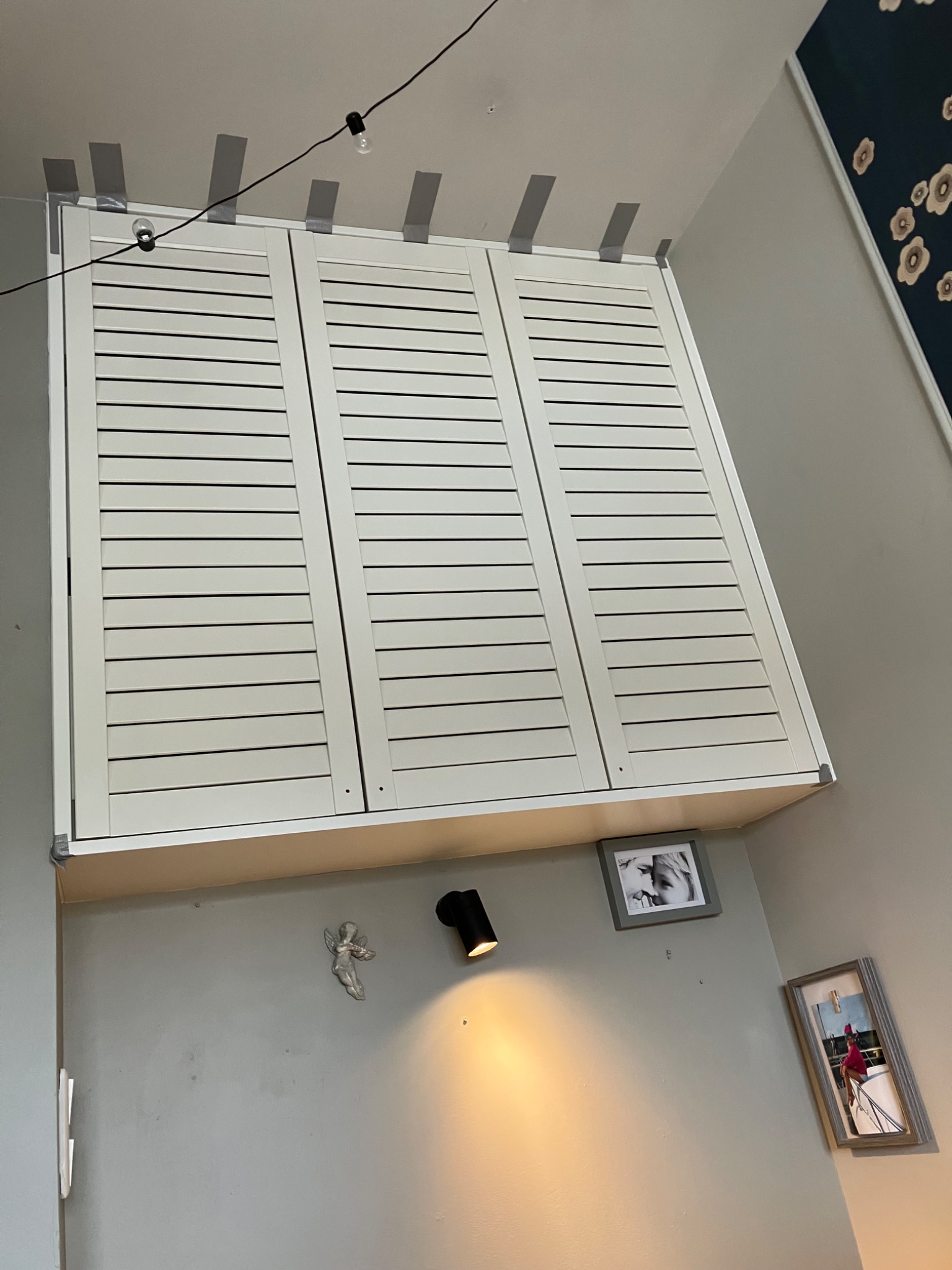 Biała szafa z żaluzjowymi drzwiami, zamontowana pod skosem na suficie, zabezpieczona taśmą malarską. Na ścianie dekoracje i oświetlenie punktowe.