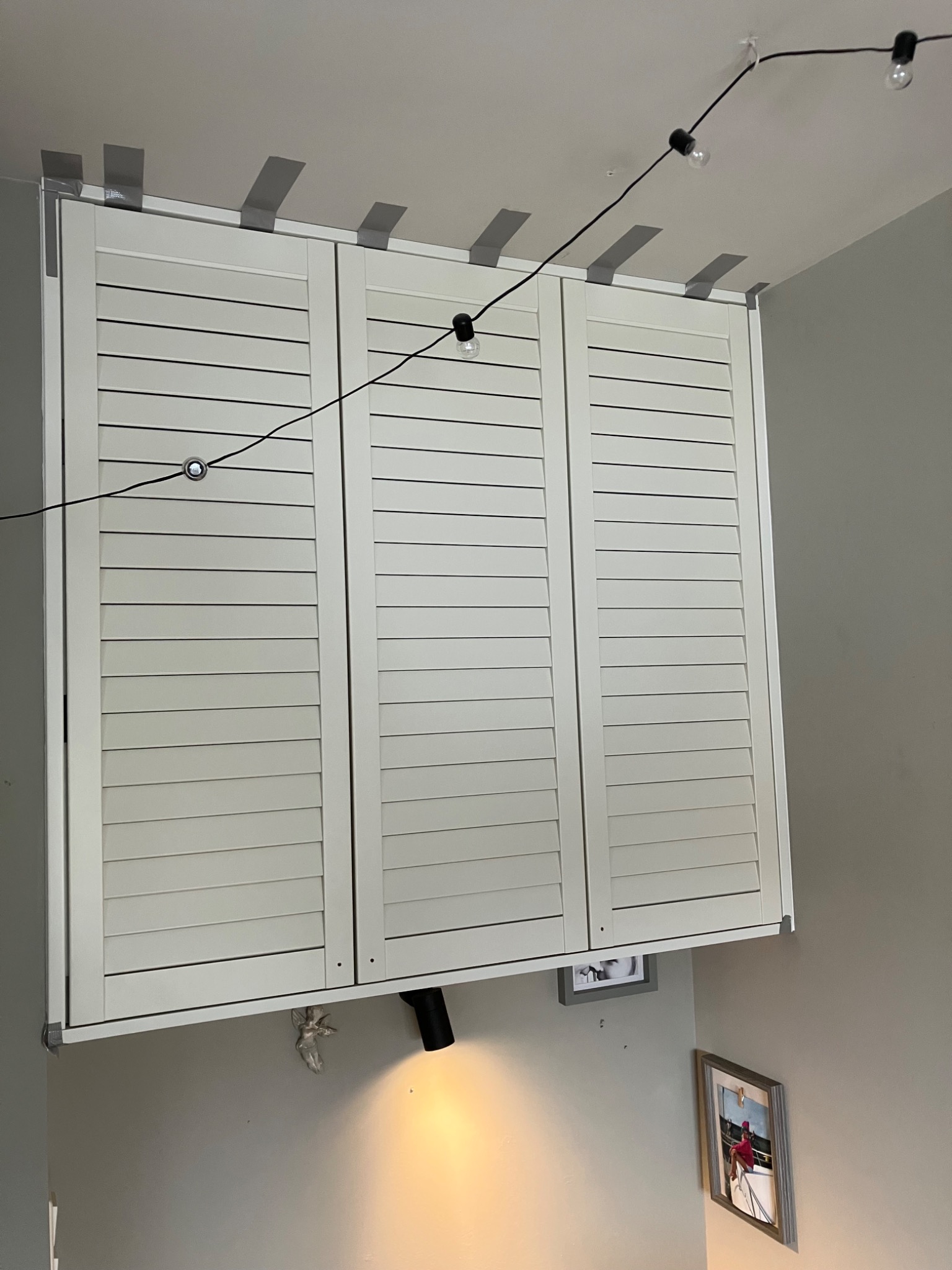 Trzydrzwiowa szafa z żaluzjowymi frontami, zamontowana pod sufitem, zabezpieczona taśmą malarską. Widoczne oświetlenie i dekoracje na ścianie.