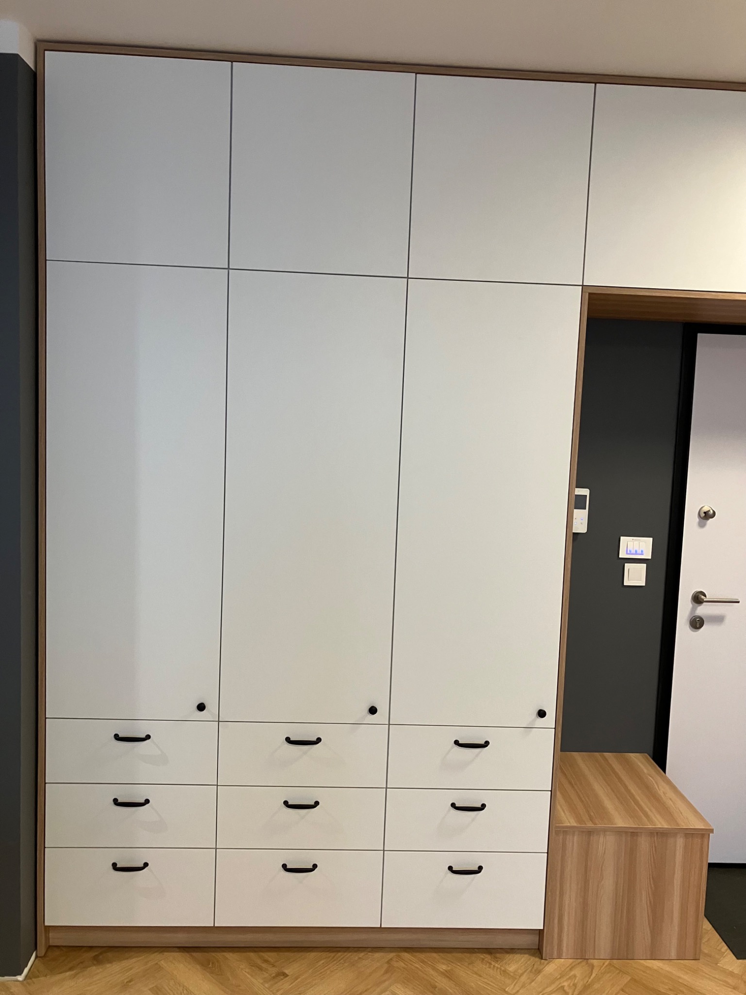 Biała szafa wnękowa z szufladami i drewnianym siedziskiem przy wejściu, minimalistyczny design, czarne uchwyty, parkiet na podłodze.